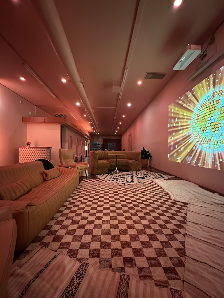 A spacious lounge area with pinkish lighting, featuring a checkered carpet, several sofas, and a large colorful abstract projection on the wall.