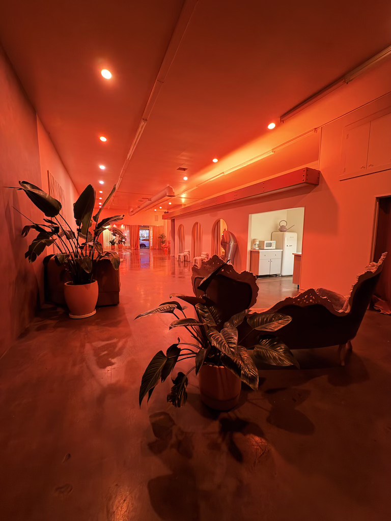 A cozy lounge area illuminated with red lighting, featuring potted plants, vintage-style armchairs, and a wall with arched mirrors; a small kitchenette with appliances is visible in the background.