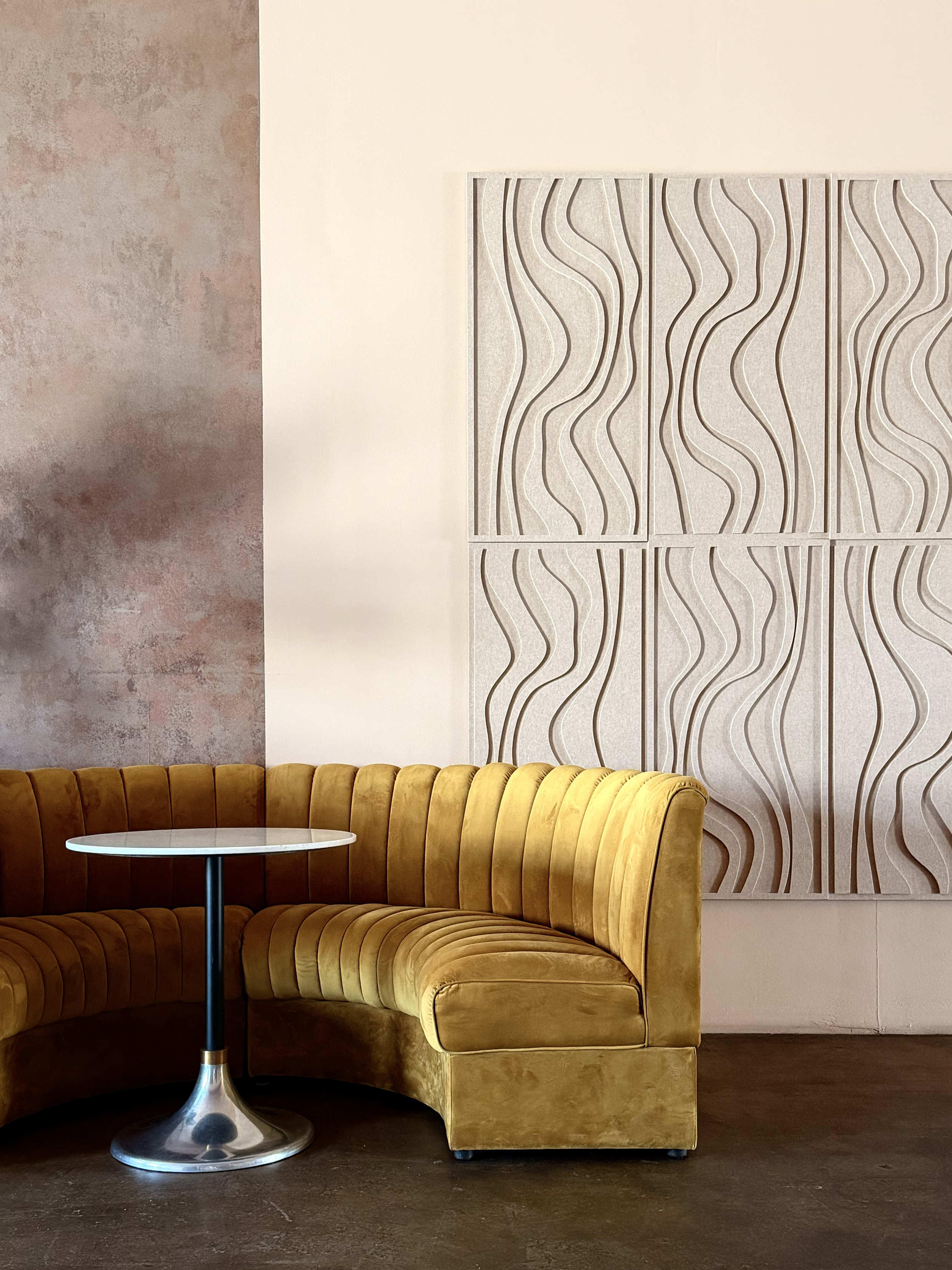 A mustard yellow curved velvet booth seat with black legs, a white round table with a metallic base, a textured brown wall on the left, a smooth white wall in the middle, and a large cream-colored abstract wall art with wavy line patterns on the righ