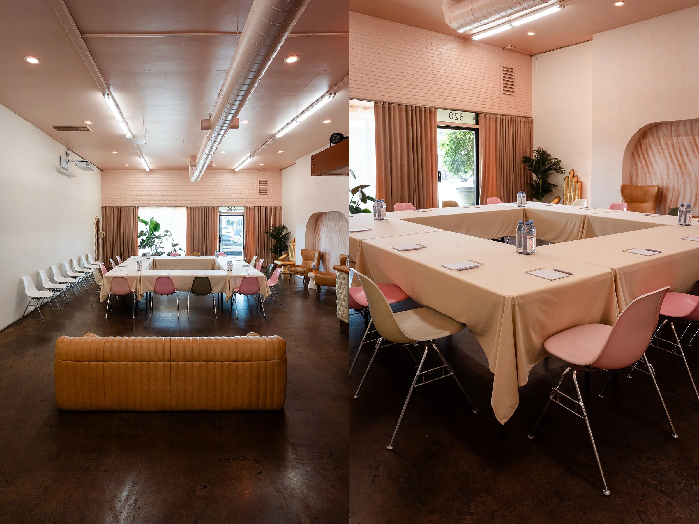 Interior of a meeting room with tables arranged in a square, beige tablecloths, and pink and white chairs. There are bottled waters on the tables, large windows with curtains, potted plants, and a mix of modern ceiling lights and exposed ductwork. Gr