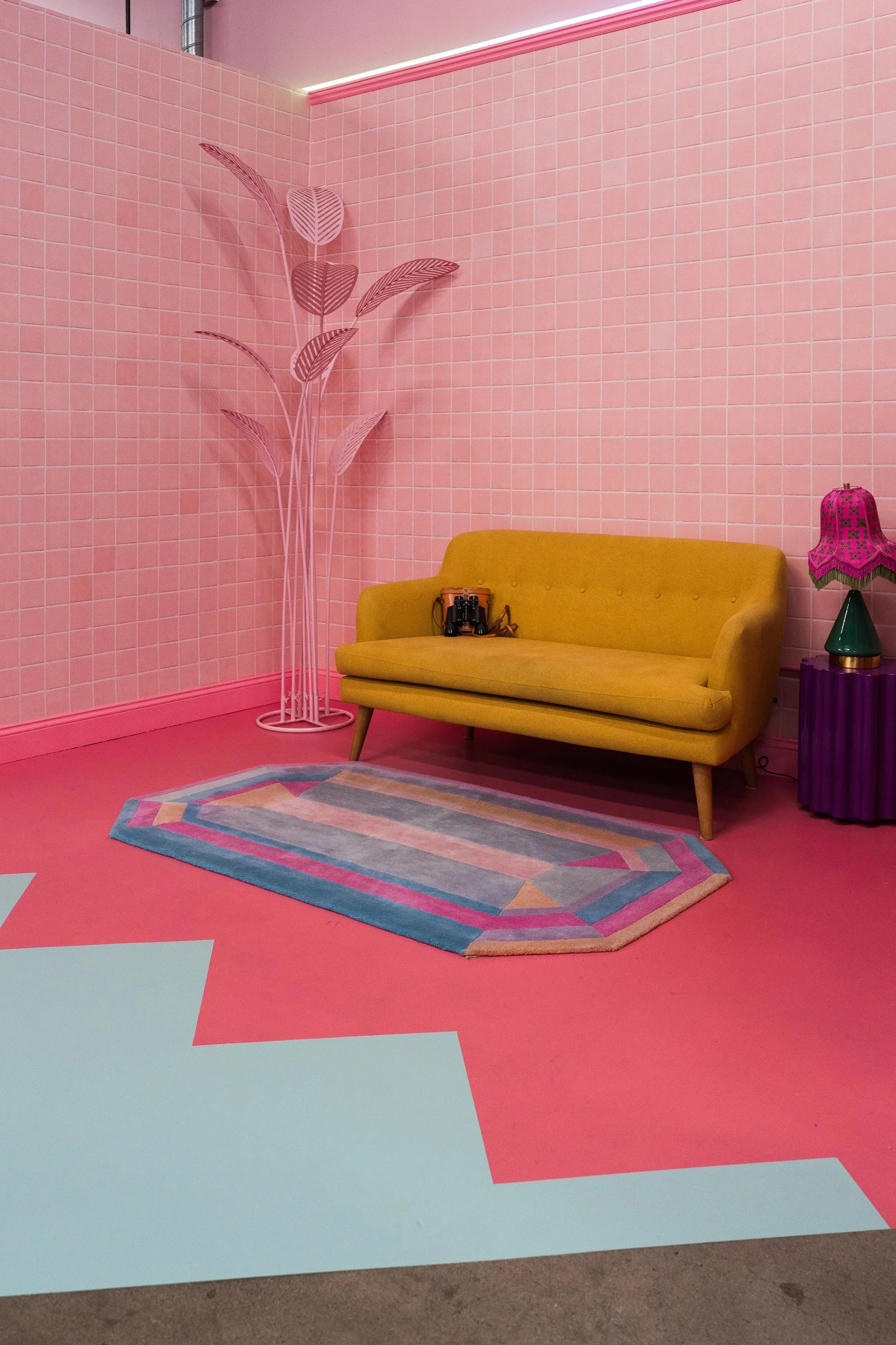 A room with pink tiled walls and pink floor. There is a yellow sofa with a pair of binoculars on it. To the right, there is a purple side table with a pink lampshade. A pastel-colored geometric rug is on the floor. In the corner, there is a tall, pink, metal plant stand with decorative leaves.