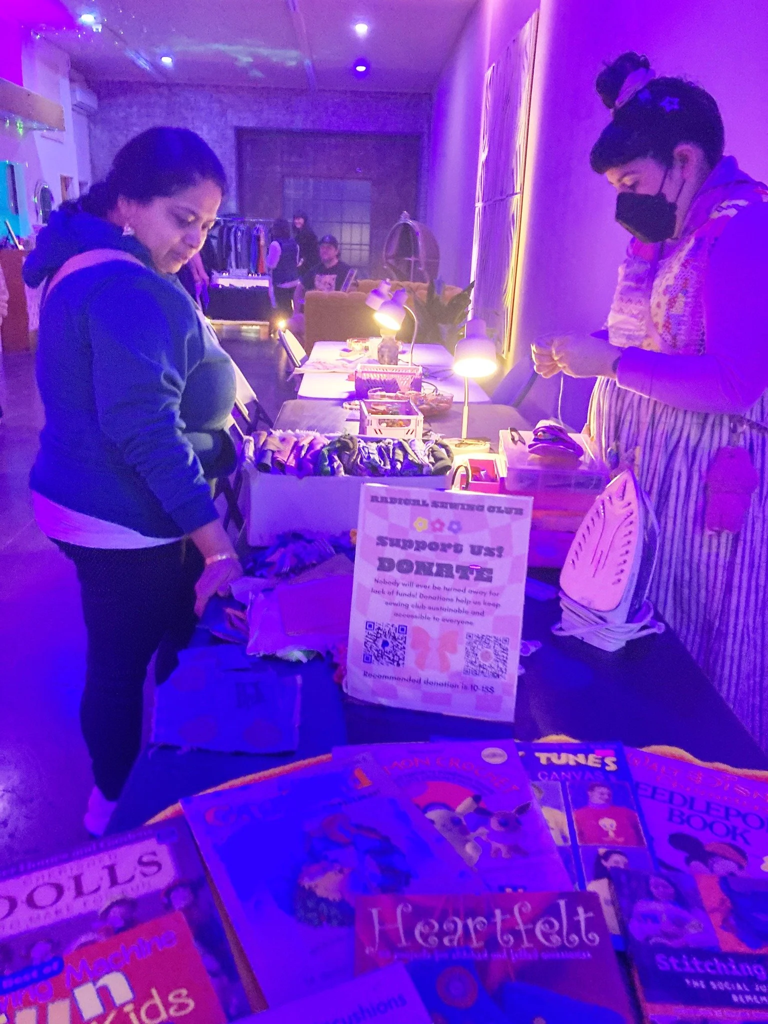 People browsing items at a table with colorful booklets and donation signs in a dimly lit room with purple lighting.