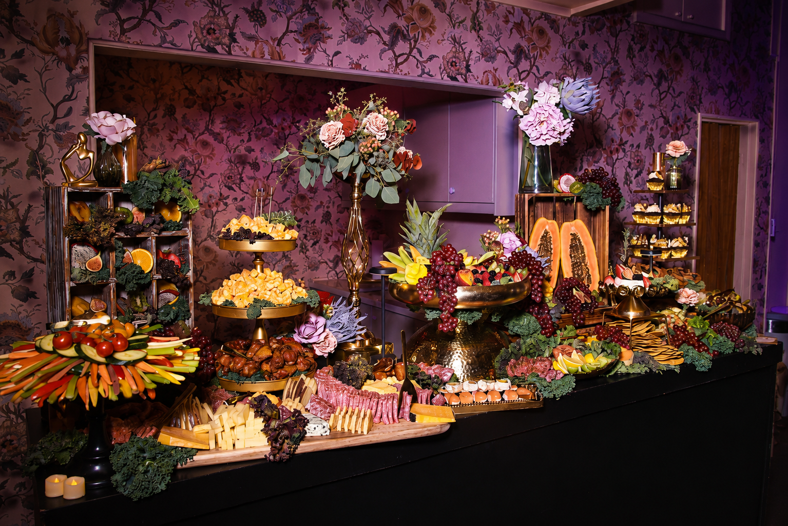 Decorative fruit and flower display with assorted fruits, vegetables, chocolates, and floral arrangements on a banquet table against a floral patterned purple wallpaper background.