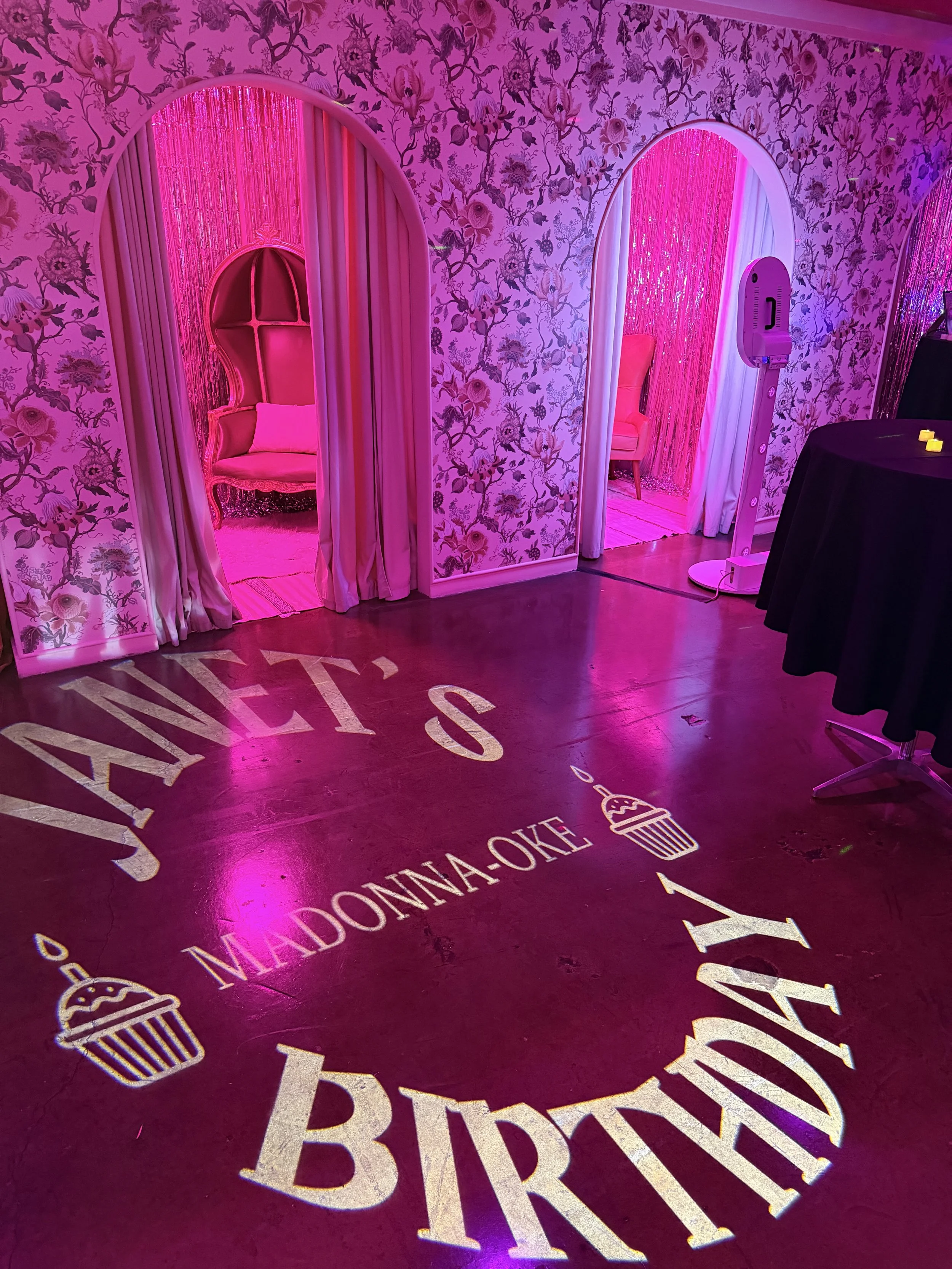 Bright pink room with floral wallpaper and pink curtains. Two pink chairs are visible through arched doorways with shimmering pink curtains. The floor has gold lettering and graphics, including the words 'MADONNA' and a dessert image.