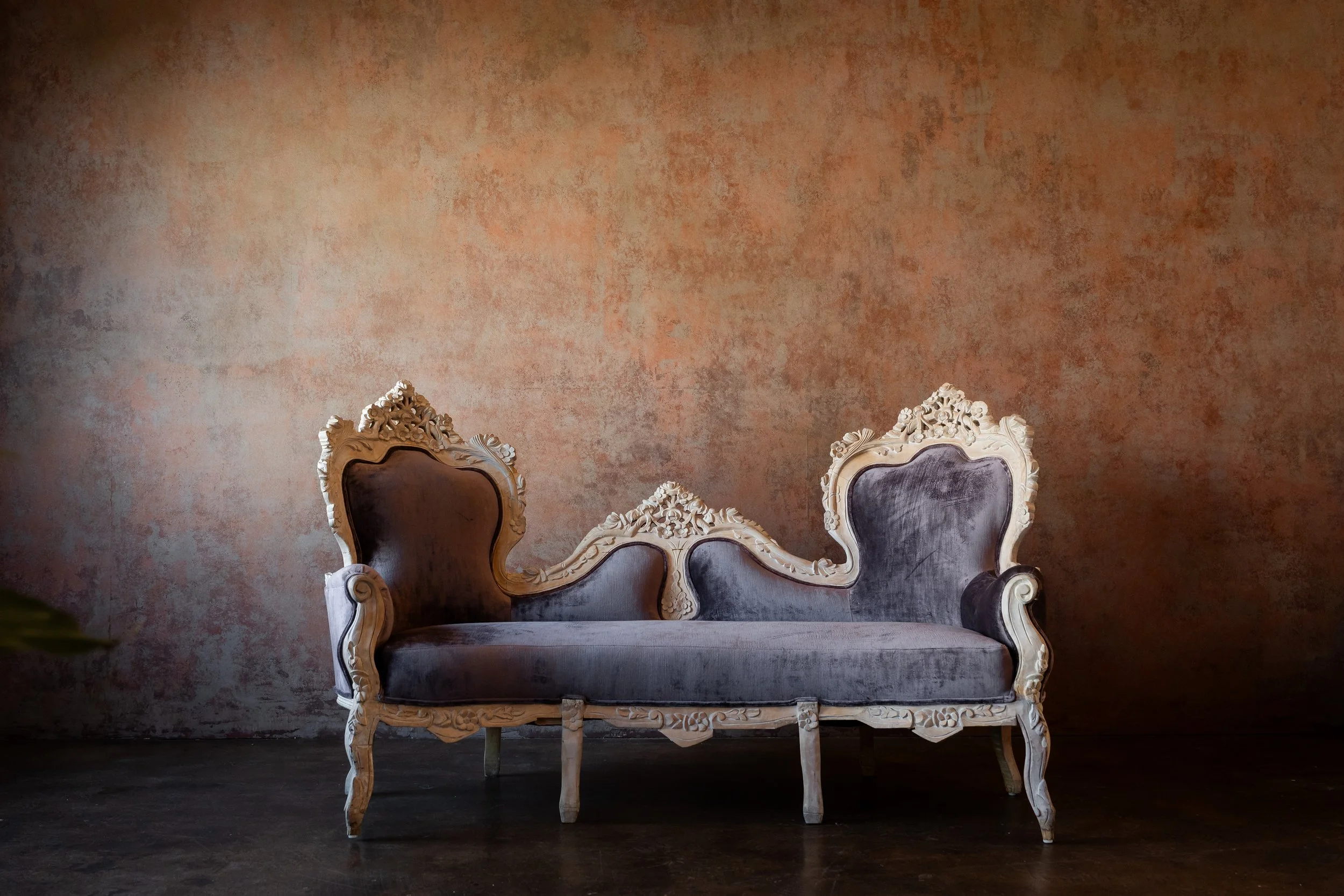 An ornate vintage sofa with carved wooden details and velvet upholstery, positioned against a textured, warm-toned wall.