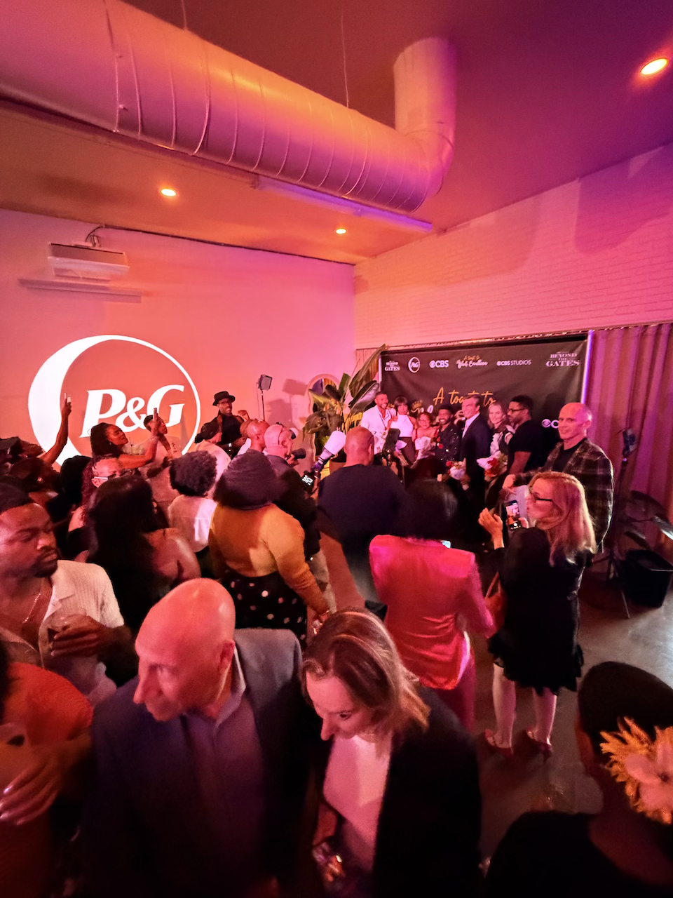 People gathered at an indoor event with a P&G logo projection on the wall, colorful lighting, and a stage with speakers and performers.