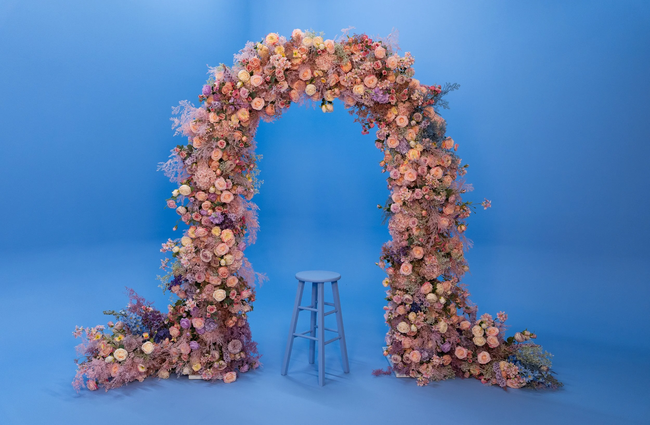 Flower arrangement arch with a stool in front against a blue background.