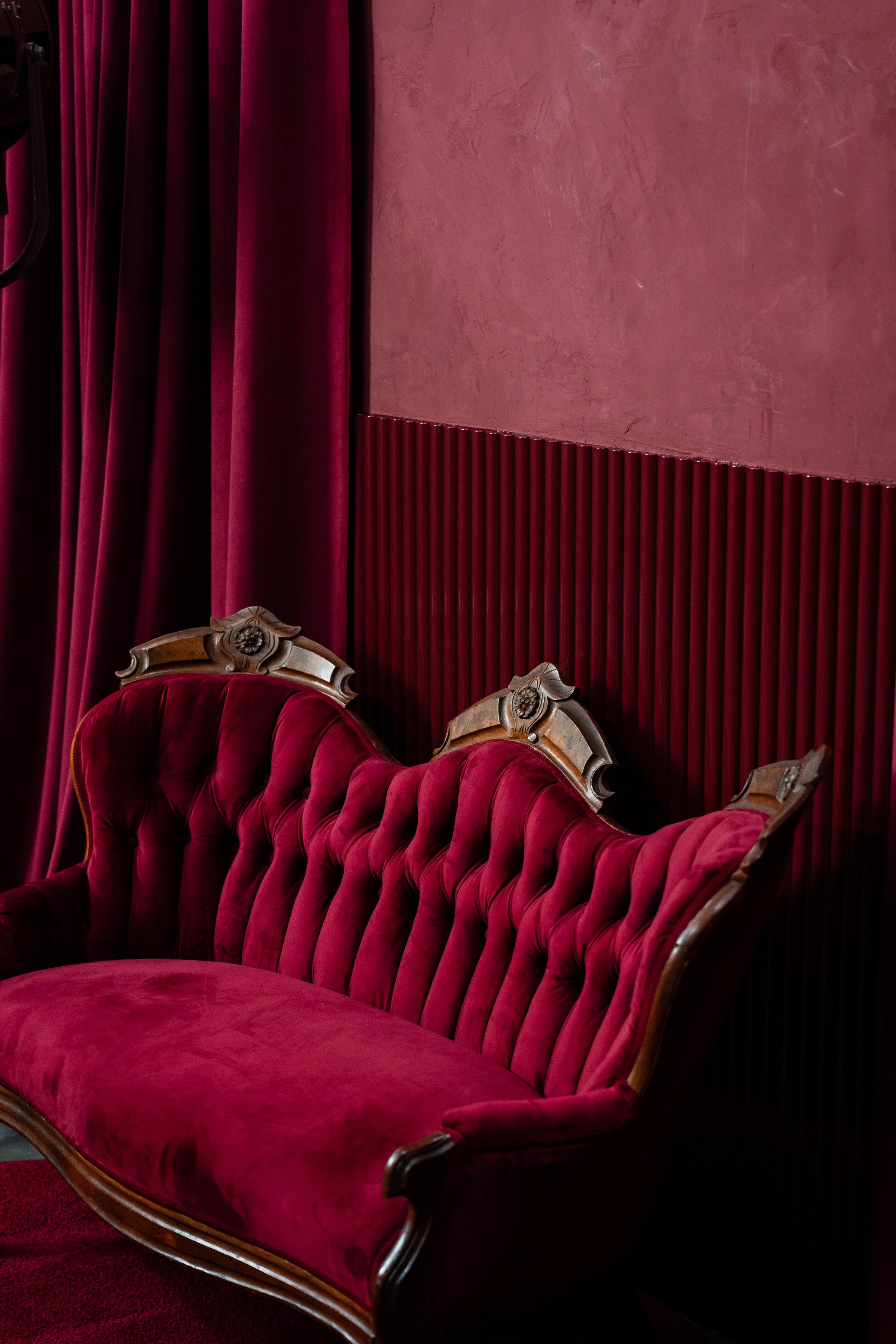 Vintage red velvet sofa with ornate wooden carvings on the backrest, next to a red curtain and wall panel in a dimly lit room.