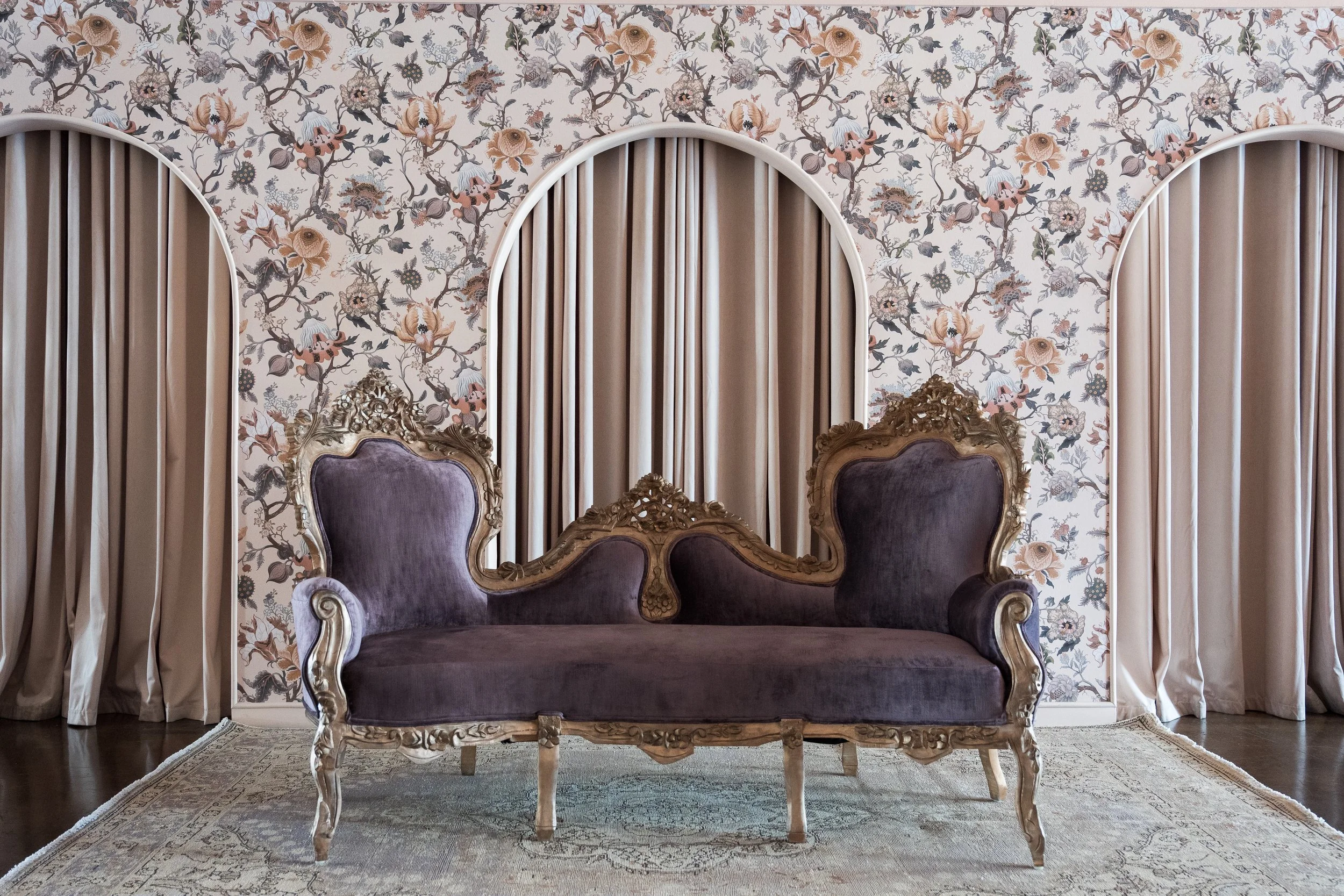 A vintage purple velvet sofa with ornate gold carved wooden frame in front of a floral wallpapered wall, flanked by beige curtains, and a patterned area rug underneath.