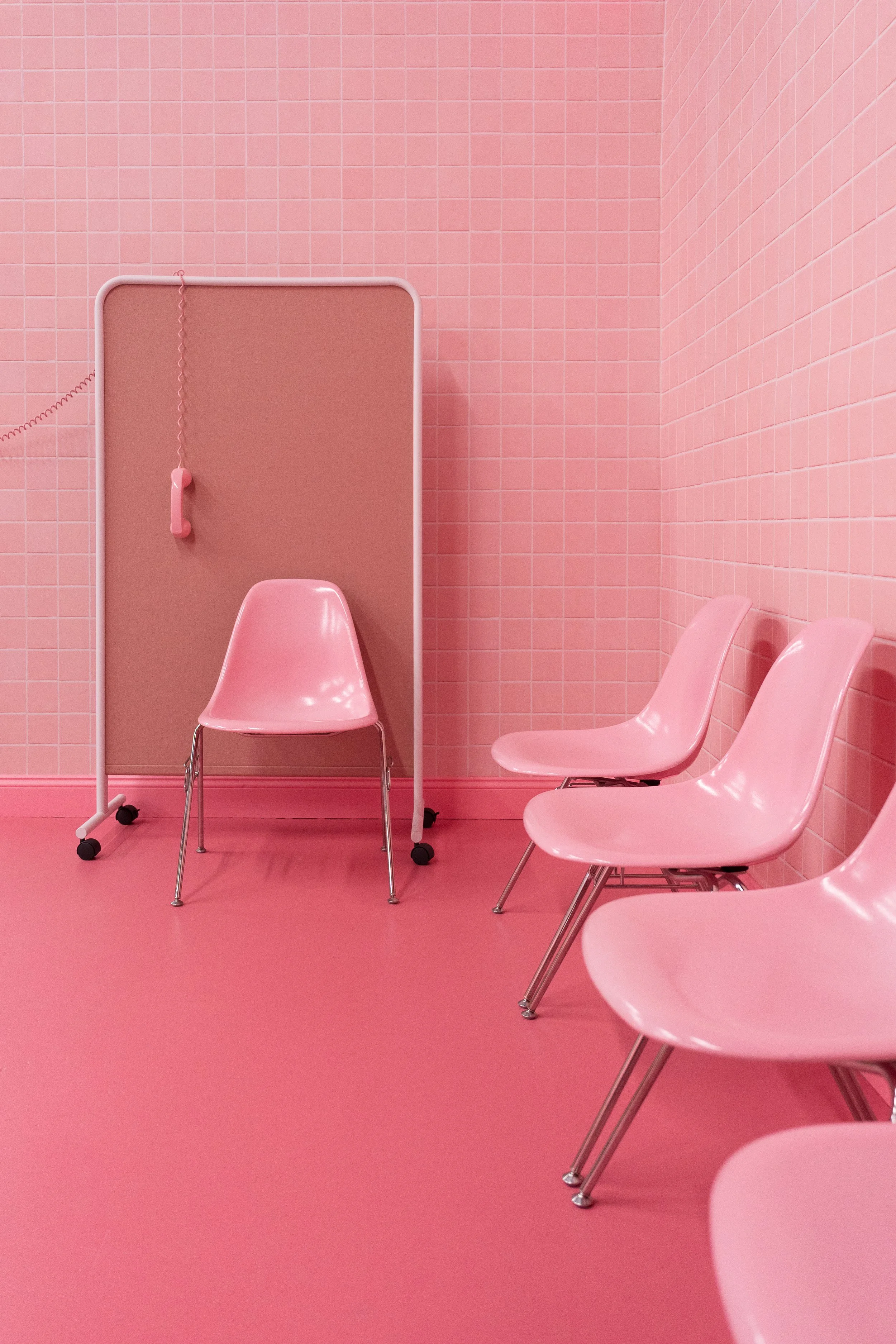 Empty pink waiting room with four pink chairs lined up against the wall, a pink towel rack, and a pink privacy partition in the corner.