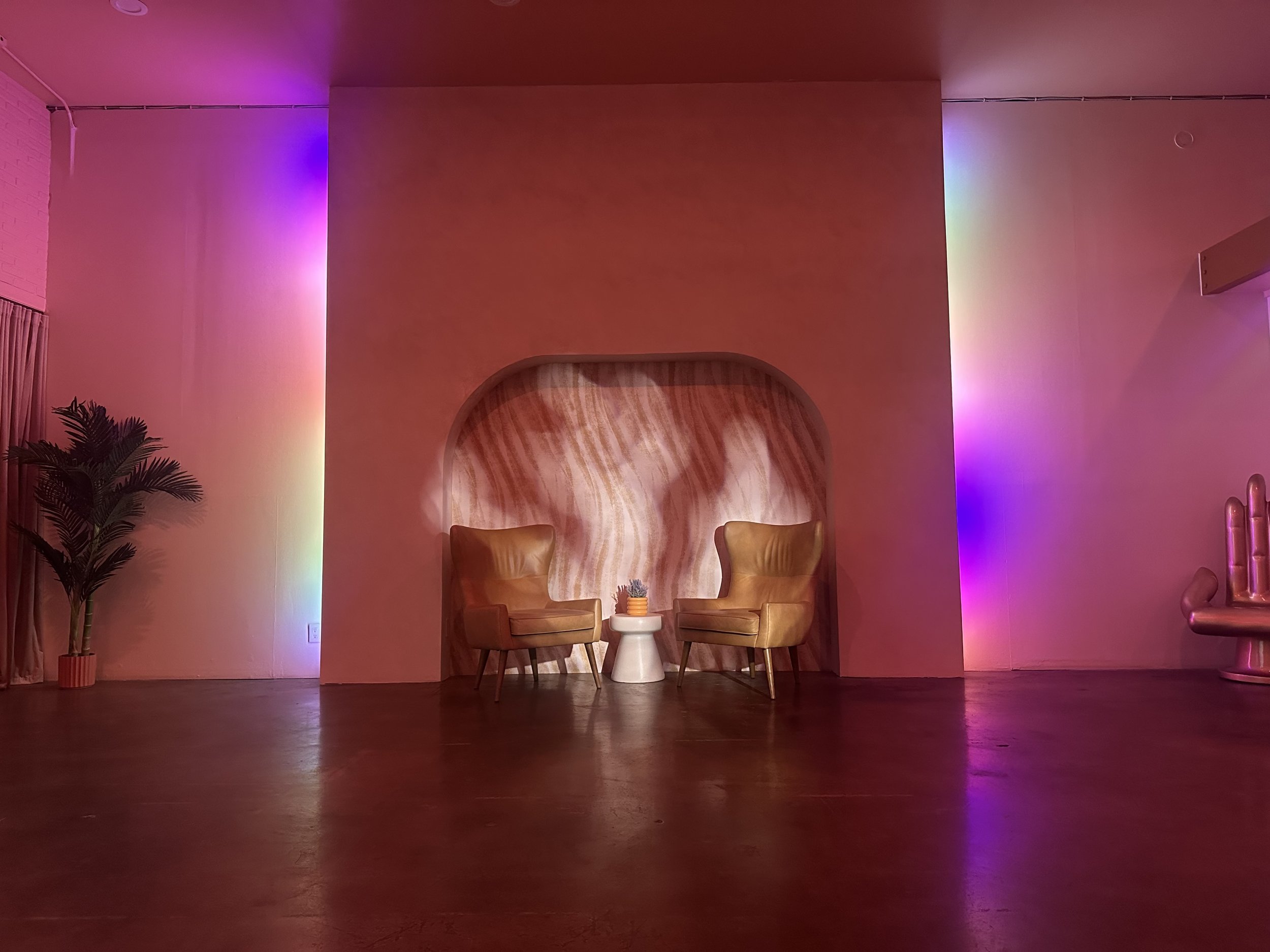 A dimly lit room with two tan armchairs facing each other in front of a wooden backdrop. There's a small round white table and a potted plant between them. The walls have colorful mood lighting creating a gradient effect from purple to yellow. A palm