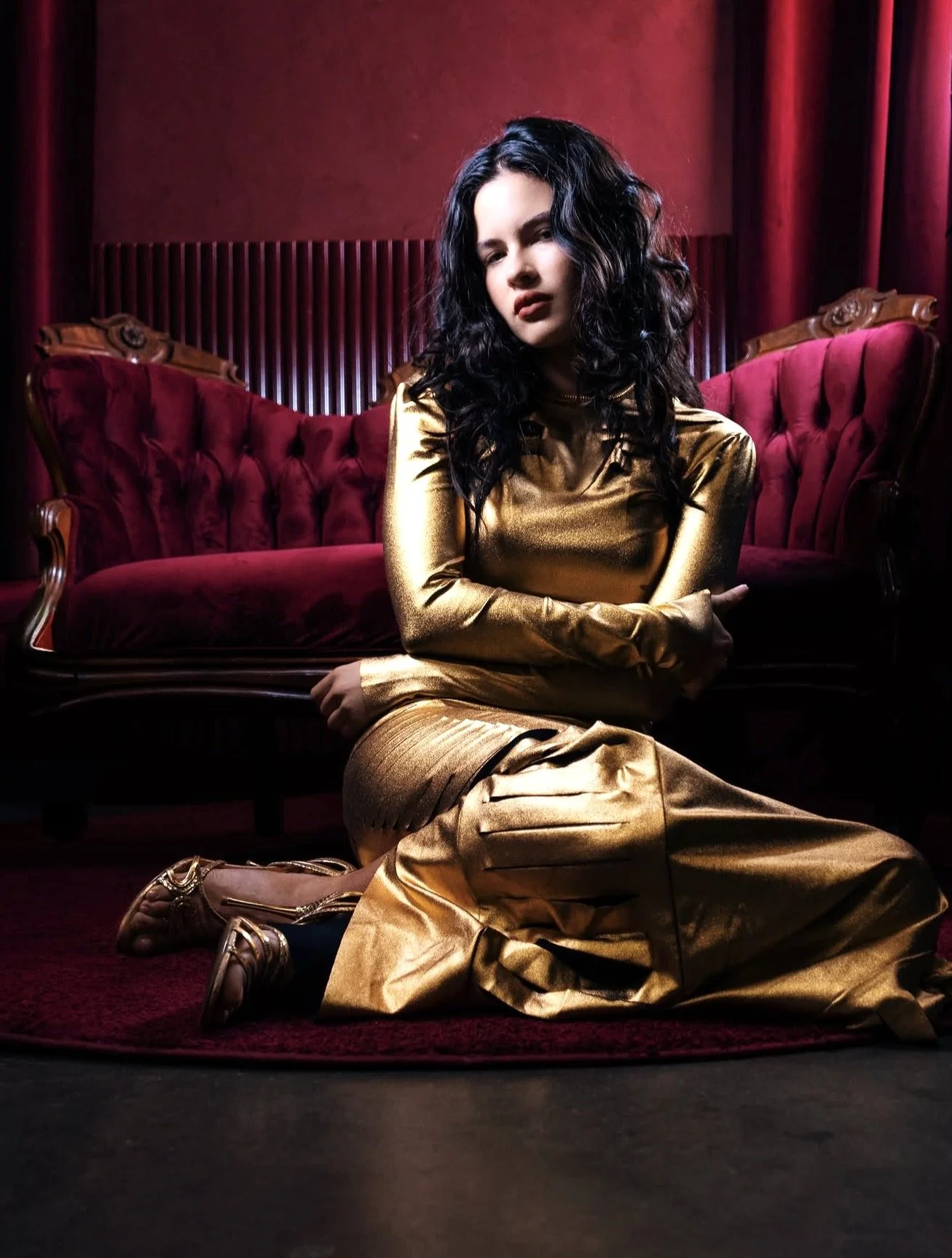 A woman with dark curly hair wearing a shiny gold outfit, sitting on a red velvet sofa with antique wooden details, in a dimly lit room with red walls and curtains.