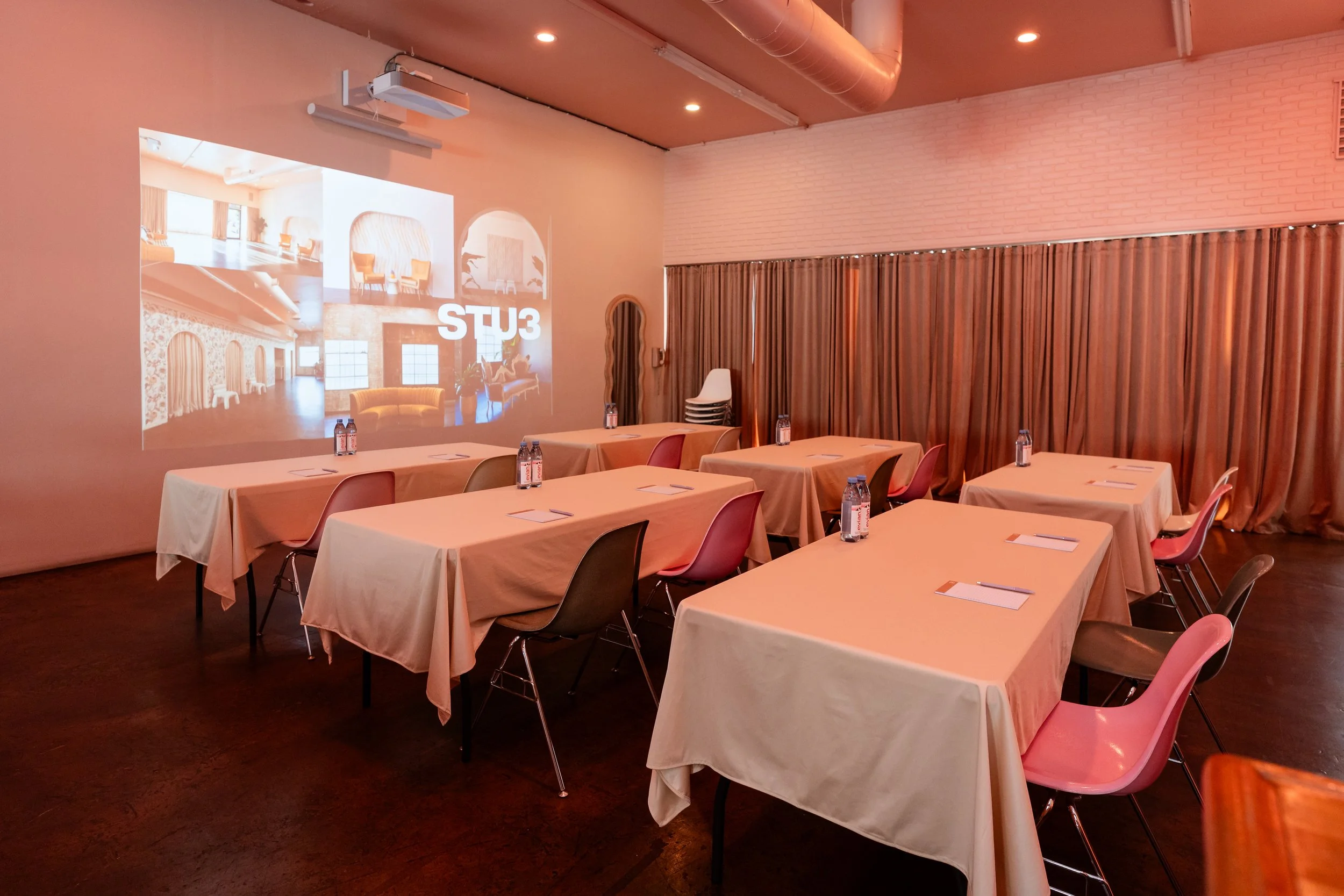 A conference room with tables covered in white tablecloths, pink chairs, water bottles, notepads, and pens, with a large projection screen displaying images of interior design ideas and seating arrangements. Great for Burbank Production Meetings, Tab