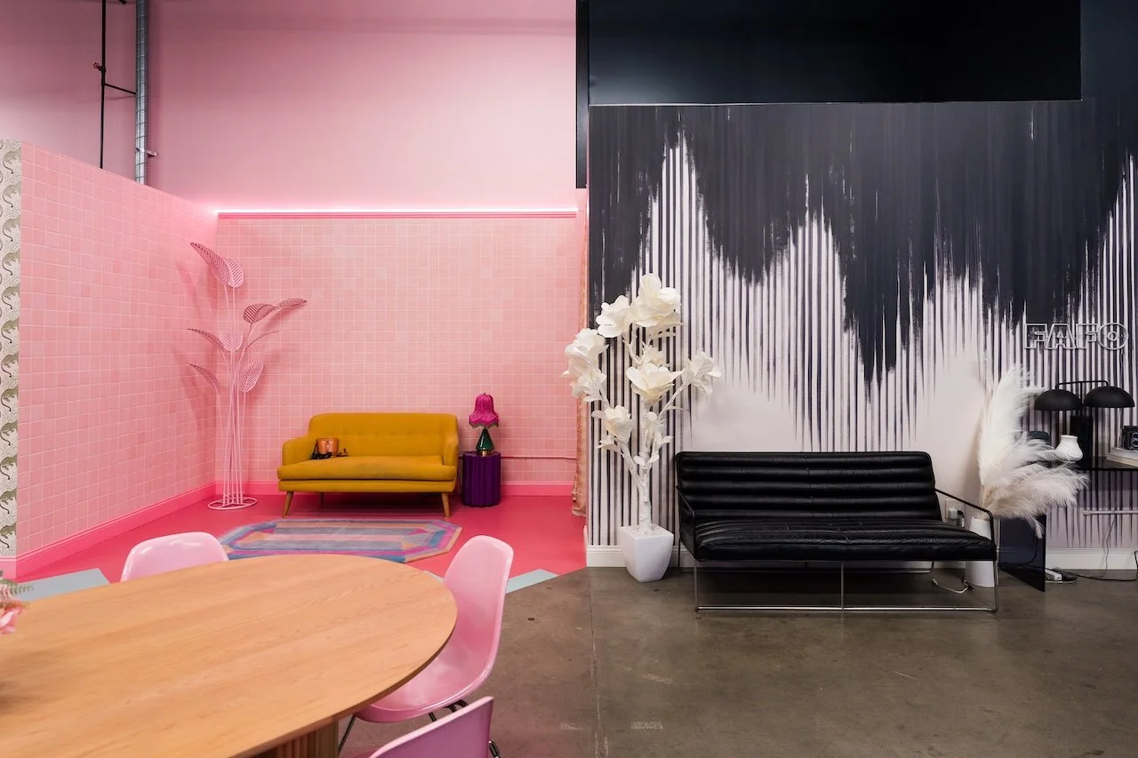 Interior of a modern room with two distinct sections: on the left, a pink corner with a yellow sofa, pink lamp, pink and blue patterned rug, pink walls, and a pink leaf-shaped decoration; on the right, a black and white area with a black leather benc