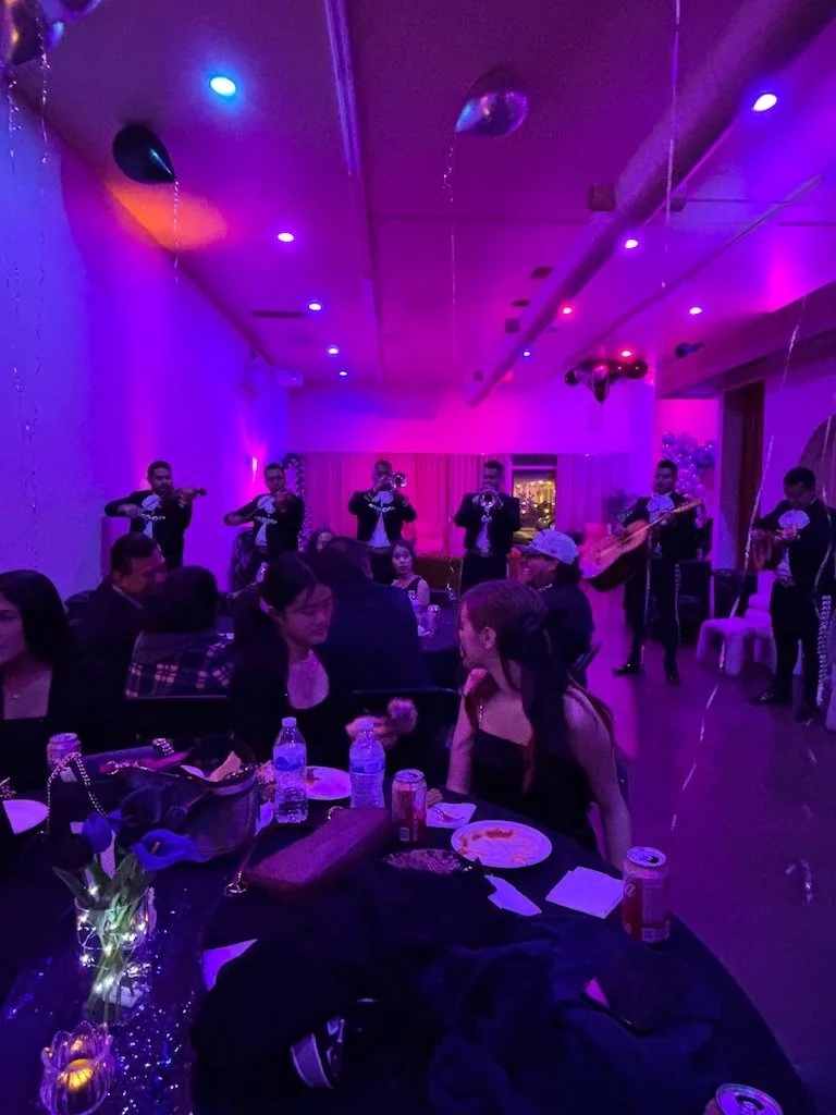 People sitting at a dinner table and enjoying live music performance in a dimly lit club with purple and blue lighting, balloons and balloons floating from the ceiling.