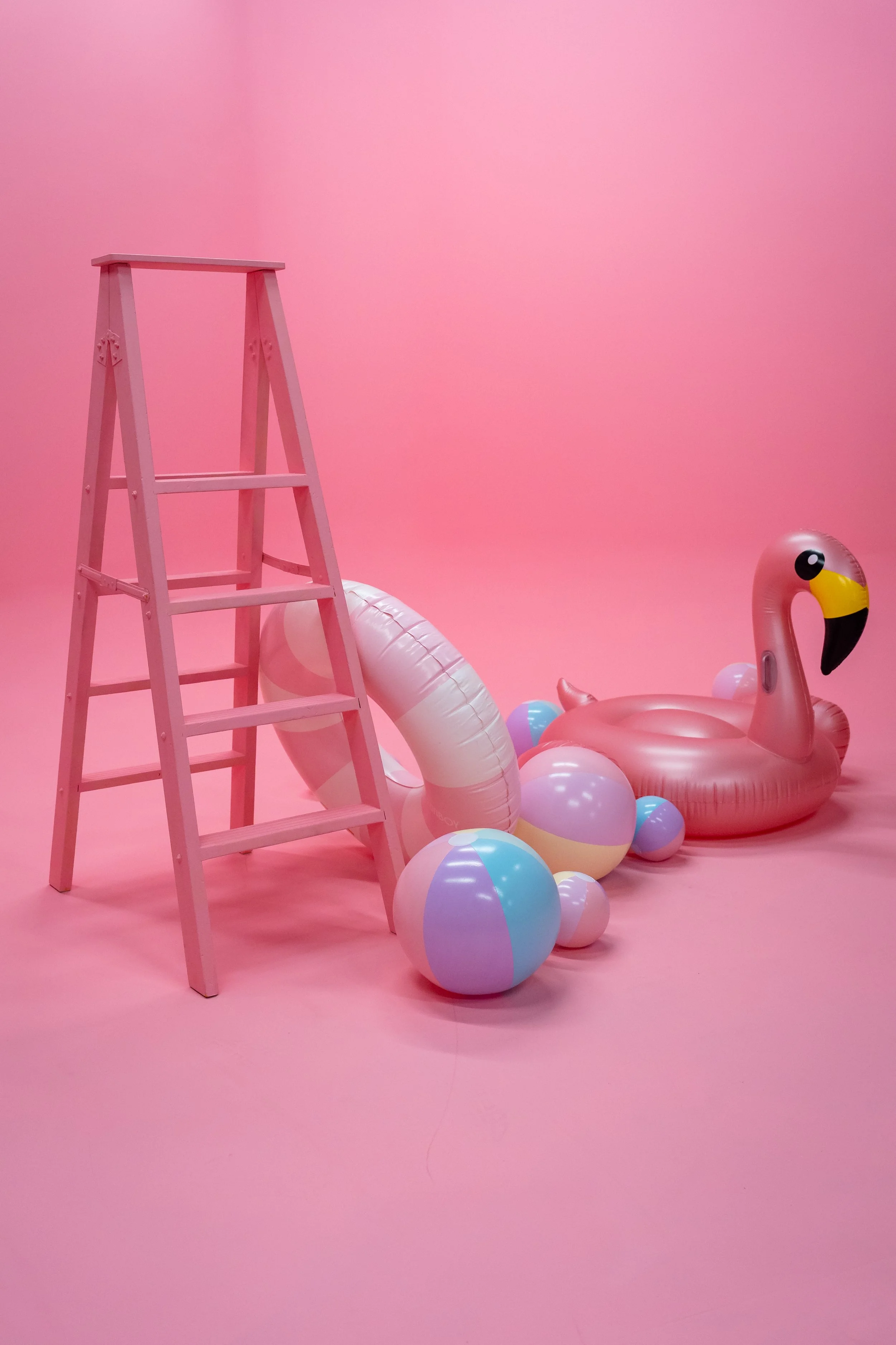 Pink inflatable flamingo pool float surrounded by colorful beach balls next to a pink step ladder on a pink background.