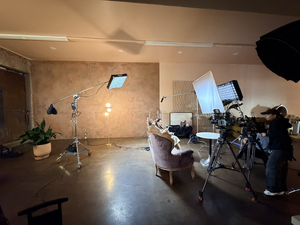 A woman sitting on a vintage chair in a dimly lit studio with professional camera equipment and lighting for filming or photography.
