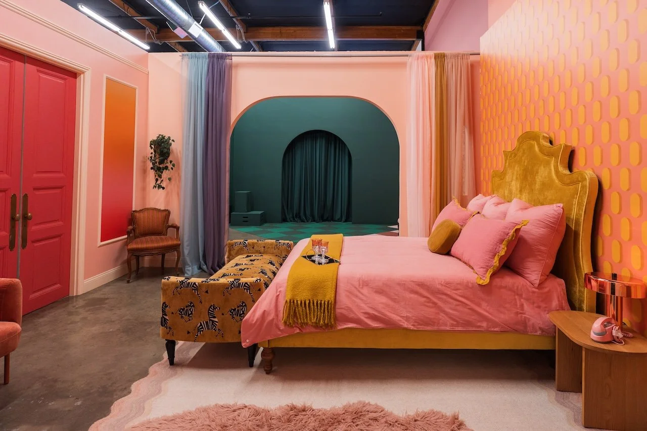 Colorful bedroom with pink bedding, gold headboard, pink and purple pillows, yellow throw blanket, pink walls with patterned accent wall, green archway with dark curtains, patterned bench, vintage chair, and pink door.
