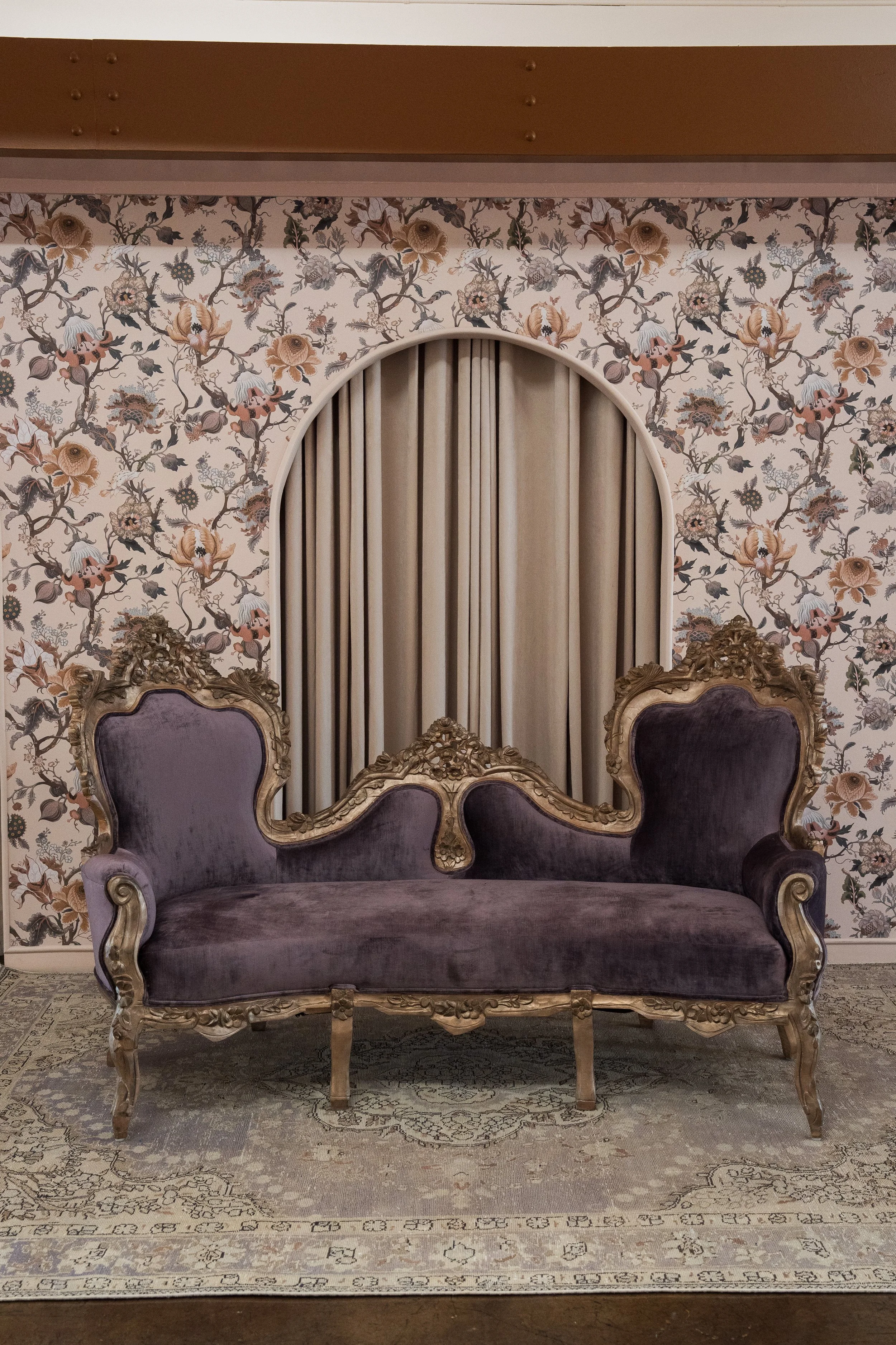 An ornate vintage sofa with purple upholstery and gold carved frame, set in front of a floral patterned wallpaper with a semi-circular window or mirror with curtains. A decorative rug covers the floor.