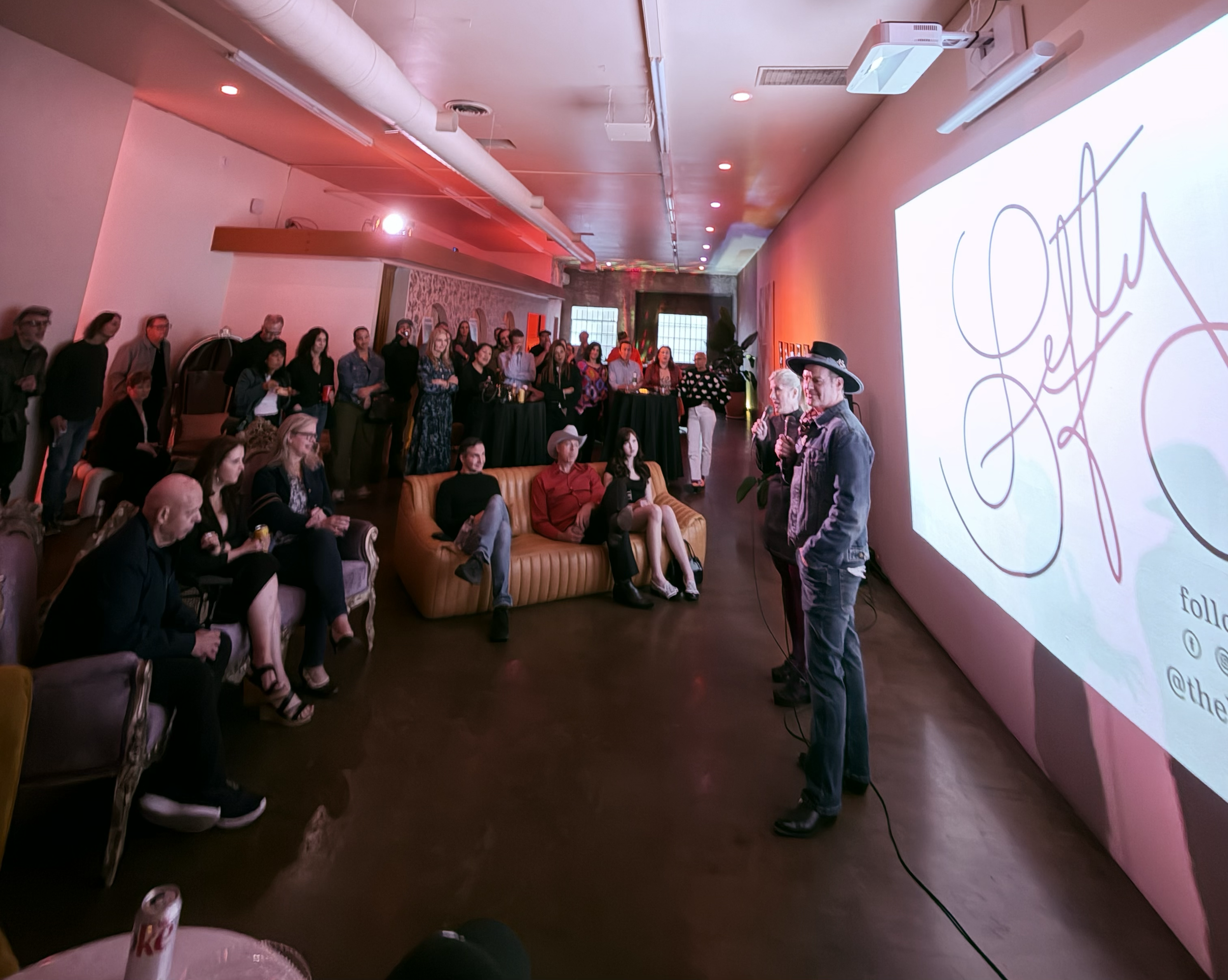 People attending an event in a dimly lit room with a large screen on the side, showing a signature or logo reading 'Etta' with social media handles. Two individuals are speaking at the front, holding microphones, with the audience seated and standing