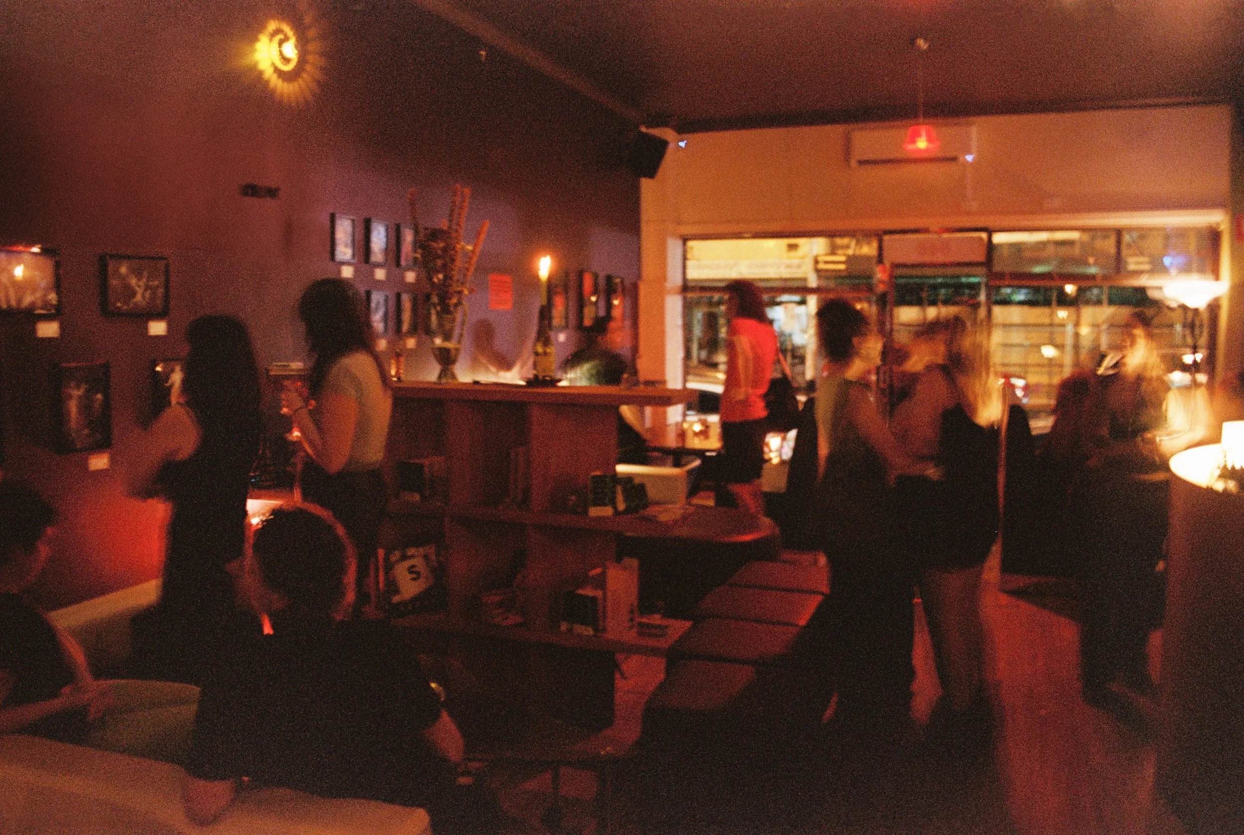 A dimly lit bar or restaurant with people socializing near a window and wall decorations, illuminated by warm reddish lighting.