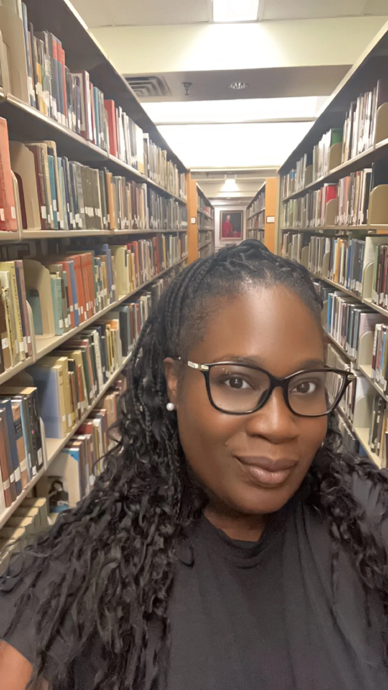 A woman with glasses and pearl earrings taking a selfie in a library aisle filled with books.