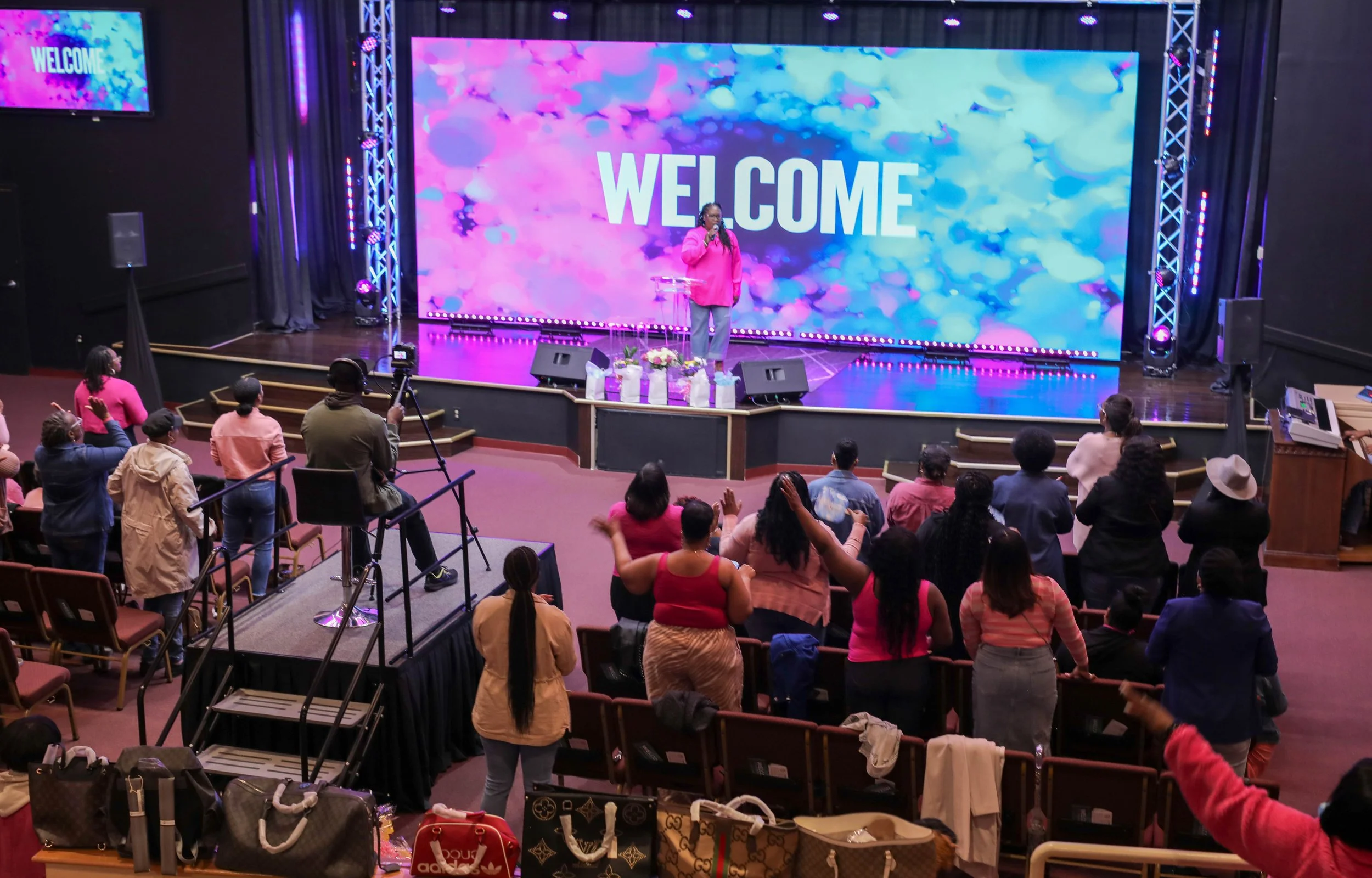 People attending a church service or event in a theater-style auditorium with a stage, where a woman in a pink top is speaking into a microphone, on a large screen behind her displaying the word "WELCOME" with colorful abstract background. Several members in the audience are standing, raising their hands, or taking photos.