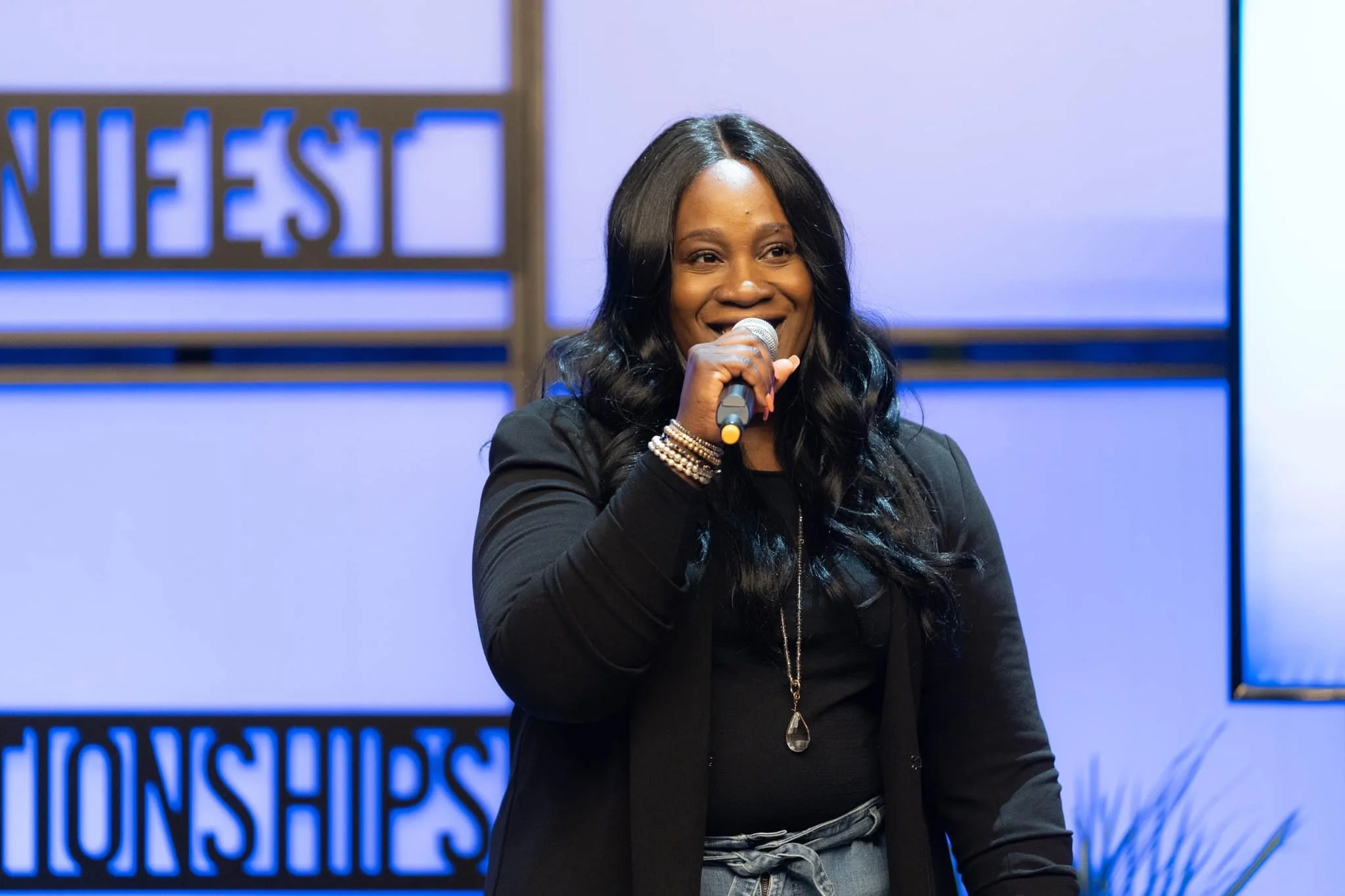Woman speaking into a microphone at an event with a blue and white background.