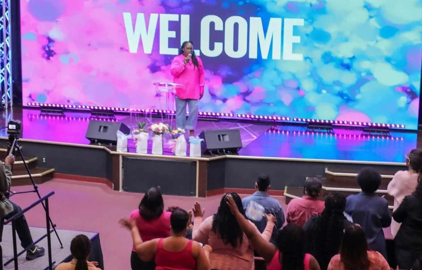 A woman in a pink blazer and gray pants speaking on stage at a welcoming event, with a large colorful screen behind her displaying the word 'WELCOME'. There are flowers and gift bags on the stage, and an audience seated and standing in front.