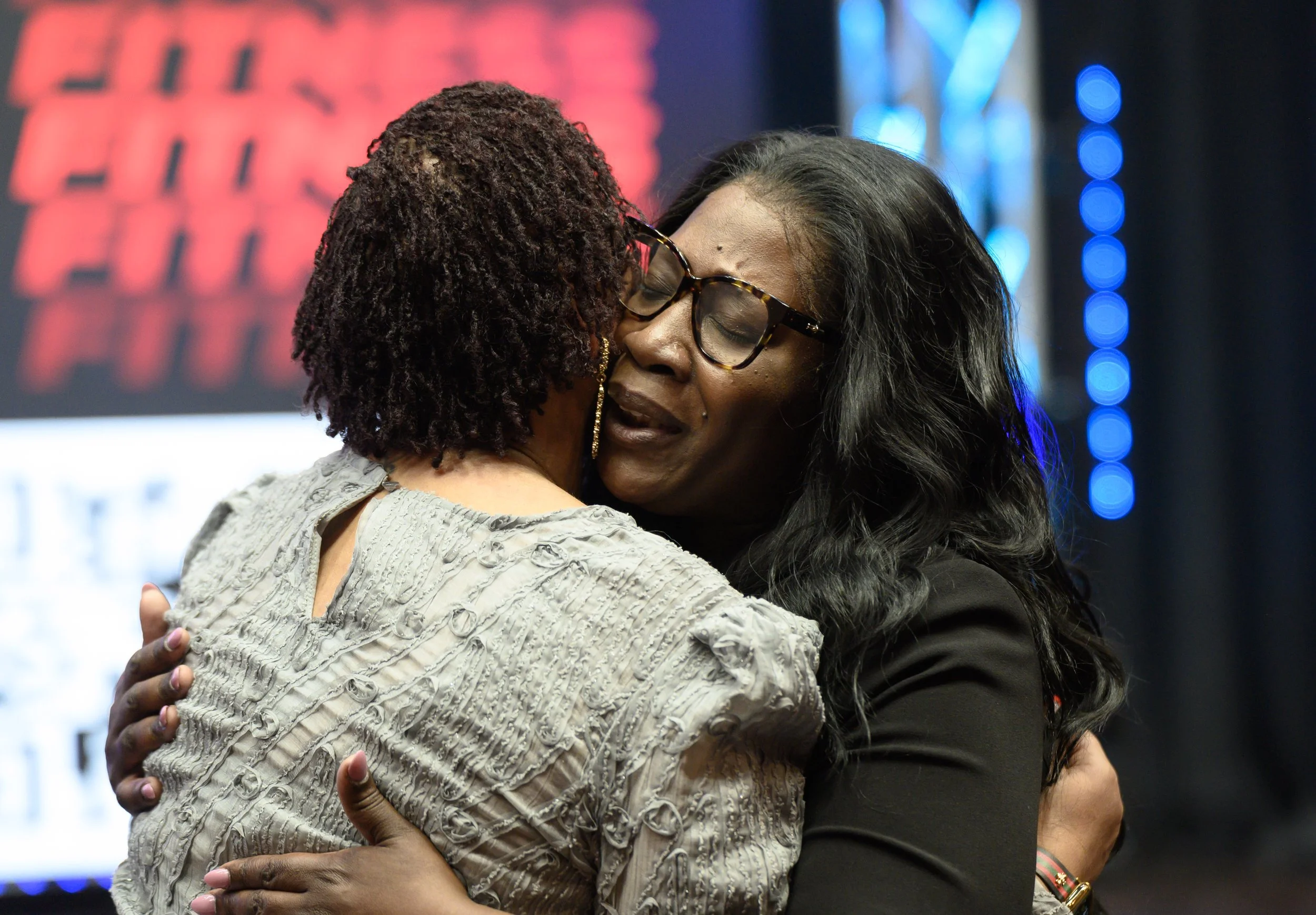 Two women are hugging each other warmly in a close embrace, one woman wearing glasses and a black jacket, the other woman with short curly hair and a textured beige sweater, in a setting with colorful and blurred background lighting.