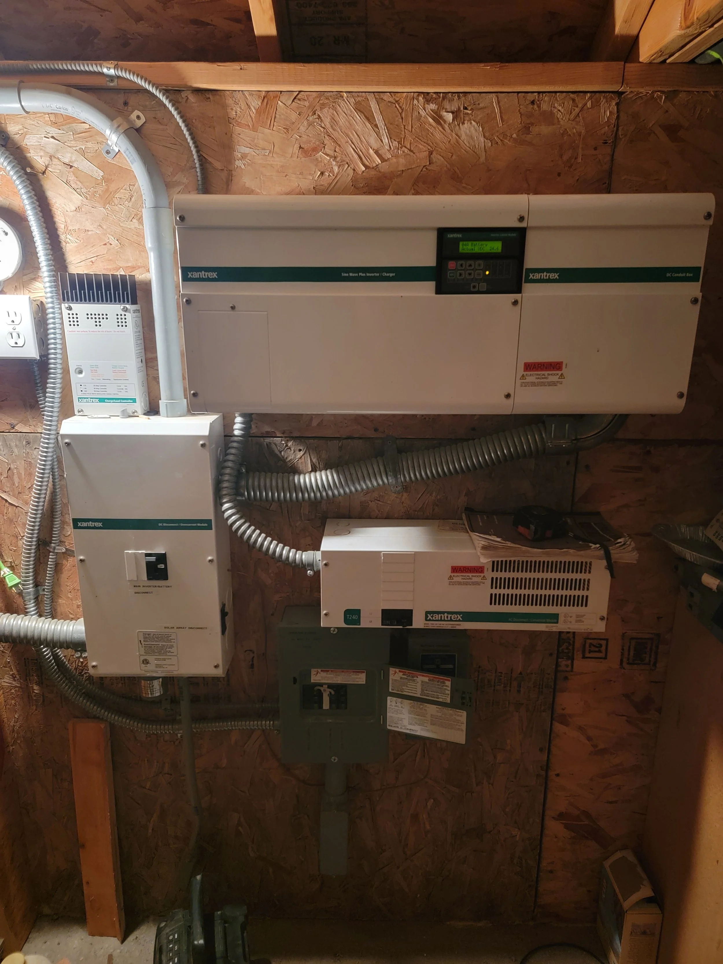 An electrical and solar power inverter setup on a wooden wall, including a solar inverter, charge controller, disconnect box, and various electrical cables.