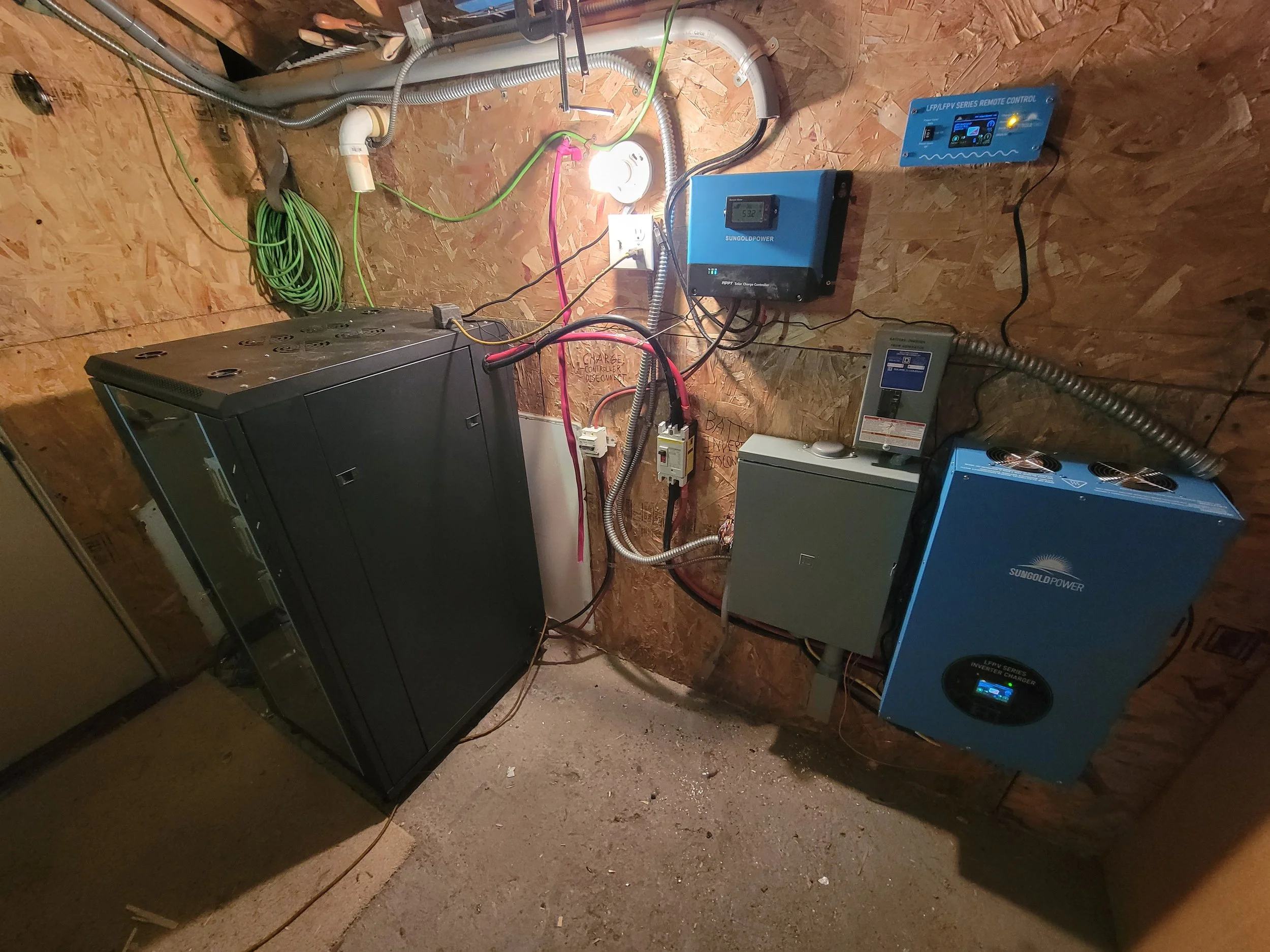 Electrical and solar power equipment installed on a wooden wall in a utility room, including inverters, controllers, and wiring for solar energy system.