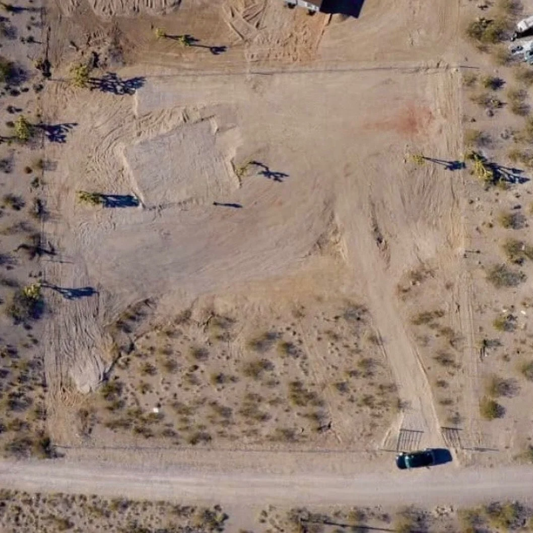 An aerial view of a barren desert area with scattered small bushes, a dirt road, and a vehicle parked at the bottom. There are shadows from sparse trees cast across the sandy terrain.
