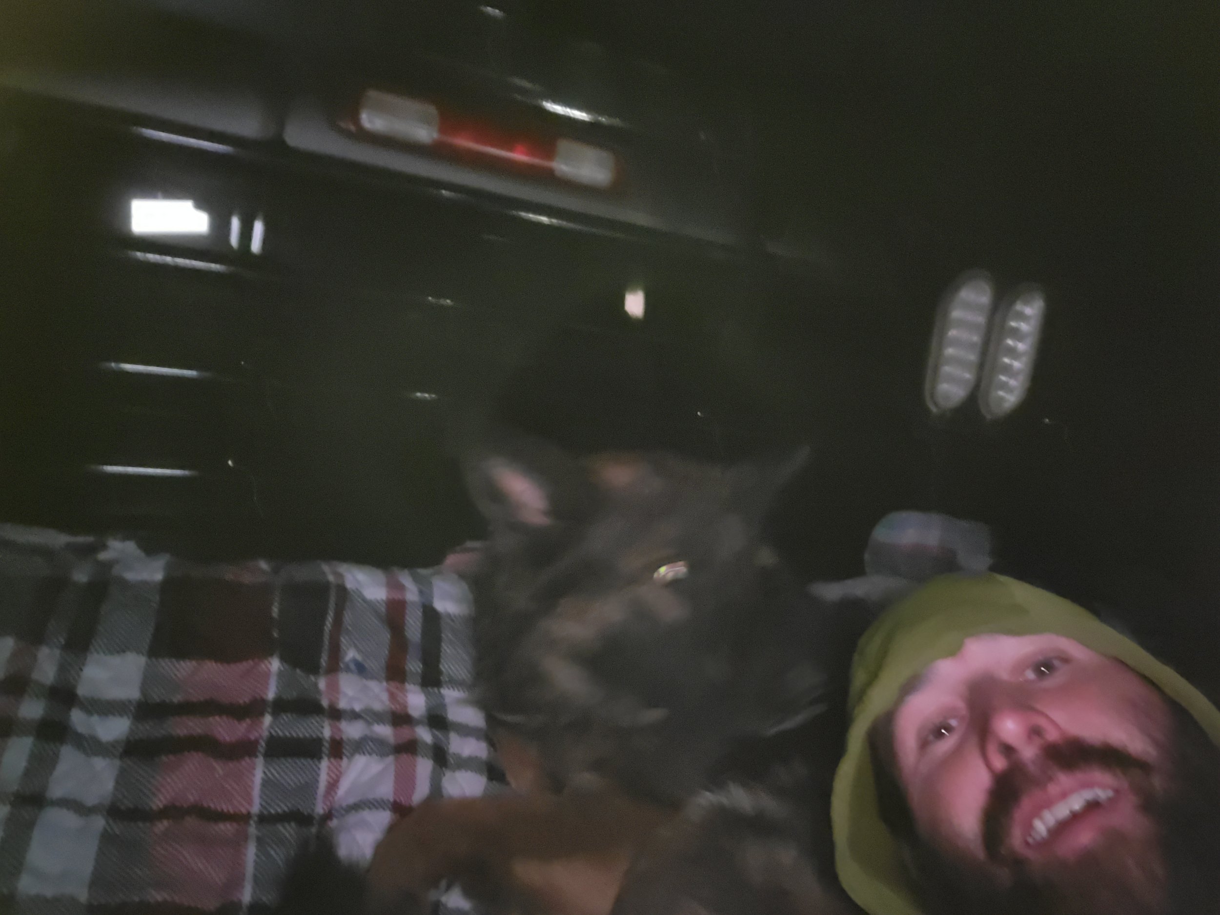 A man lying inside a vehicle, smiling with a black and tan dog resting on his chest, and a person wearing a yellow hat nearby. The vehicle interior and part of the outdoors are visible.