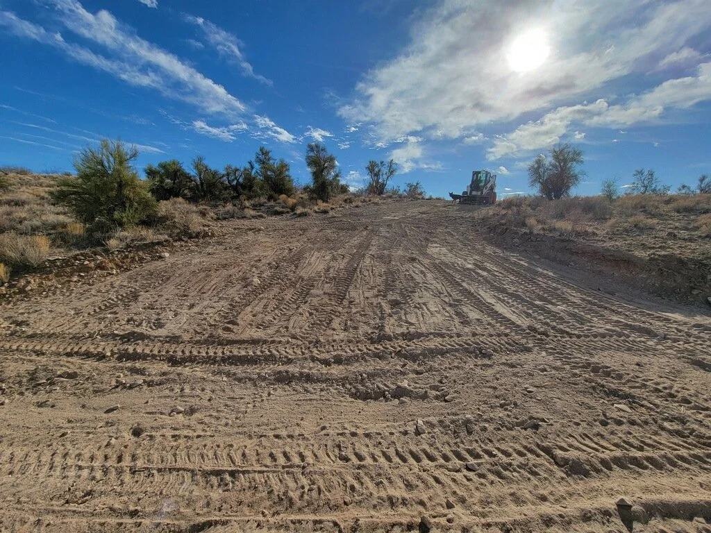 A dirt road with tire tracks, small bushes, and a construction vehicle on a hill under a partly cloudy sky with the sun.