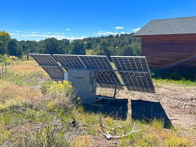 A solar panel array installed on a gray metal structure in a grassy field with a dirt path, a red barn, trees, and a blue sky with some clouds in the background.