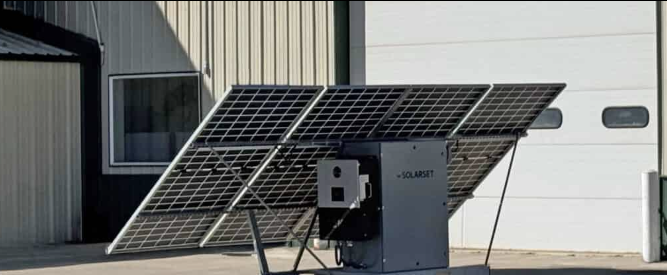 A large solar panel array installed outdoors next to a building with a white garage door and a smaller building with a window.
