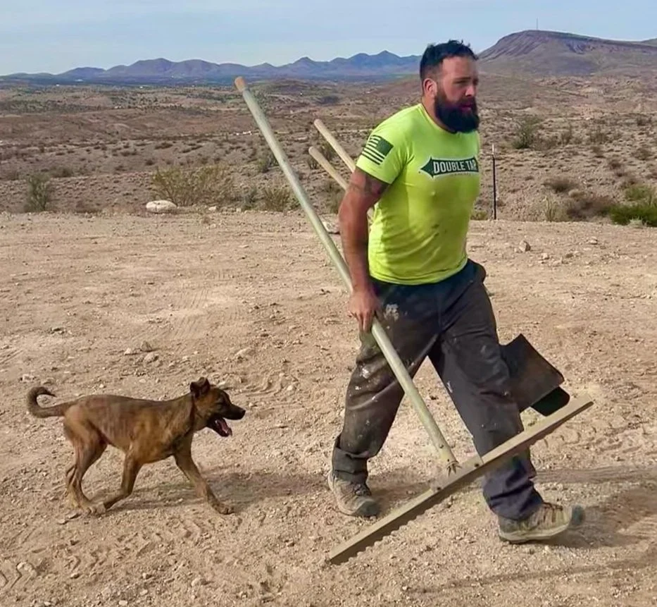 A man wearing a neon green shirt and dark pants playing mini golf in a desert area with a dog running behind him.