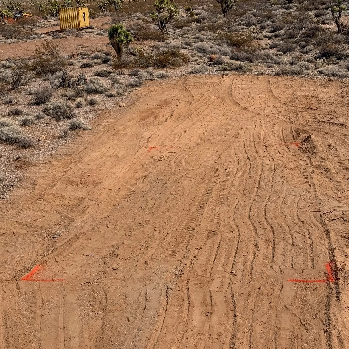 A dirt road with tire marks and orange spray paint markings on it in a desert landscape with sparse bushes and small trees. There is a yellow container or structure in the background near the horizon.