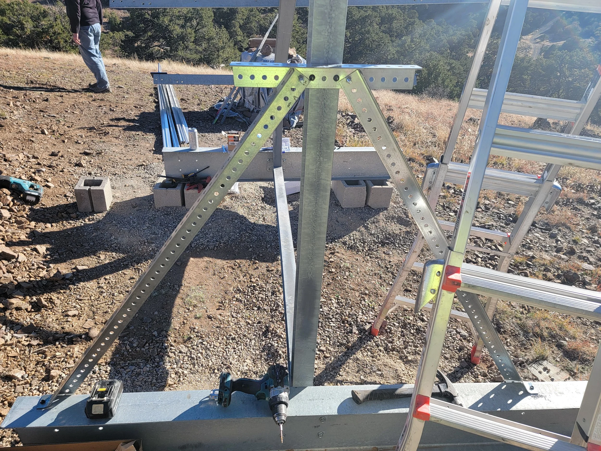 Construction setup on a hillside with ladders, metal beams, and tools, with a person standing in the background overlooking a scenic landscape.