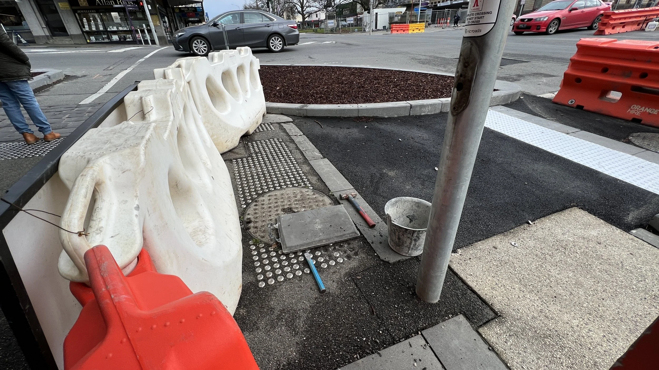 Construction site on a sidewalk with white barriers, construction tools, and a utility pole, next to a street with cars and shops as Stainless Discrete Tactile Ground Surface Indicators (TGSIs) are being recovered prior to new infrastructure work.