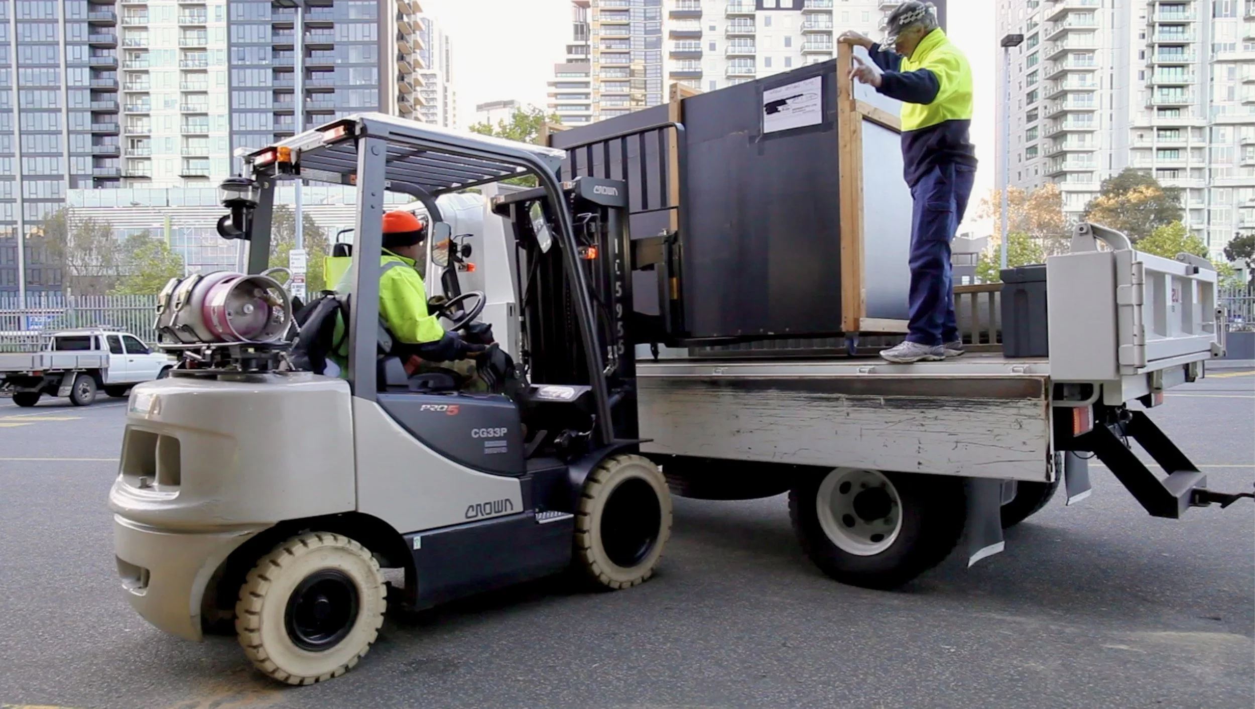 Workers loading a large black crate onto a truck using a forklift, with tall buildings and parked vehicles in the background.