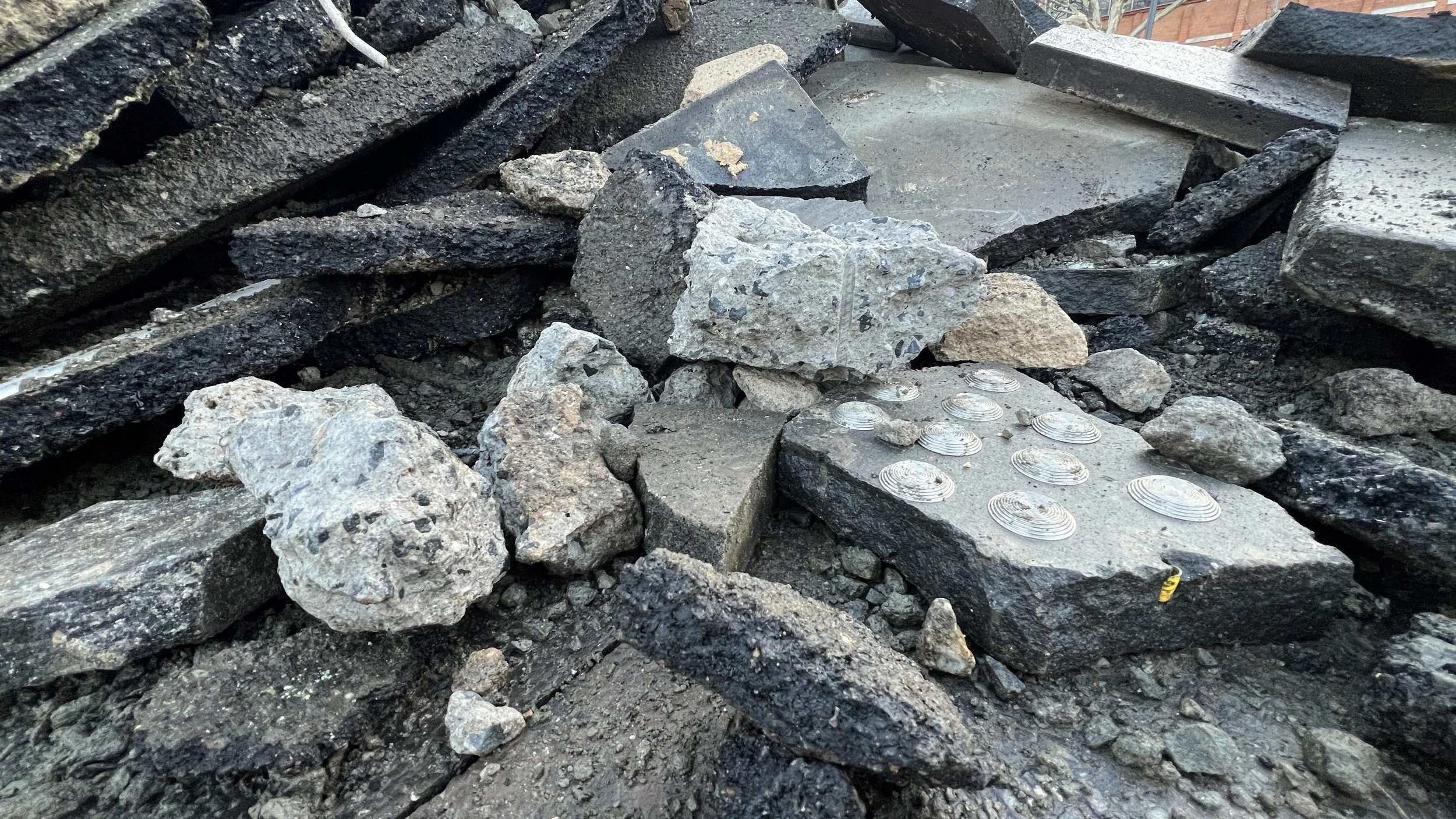 Close-up view of a construction demolition site with broken concrete and asphalt debris, some large rocks, and a section of concrete with embedded 20 year old Stainless Steel Discrete Tactile Ground Surface Indicators (TGSIs) still installed.