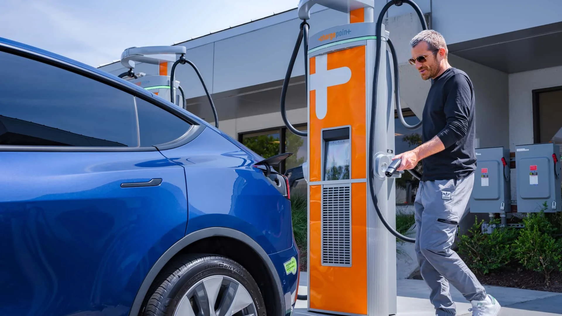 A man charging a blue electric car at an orange ChargePoint DC fast charging (DCFC) station outdoors.