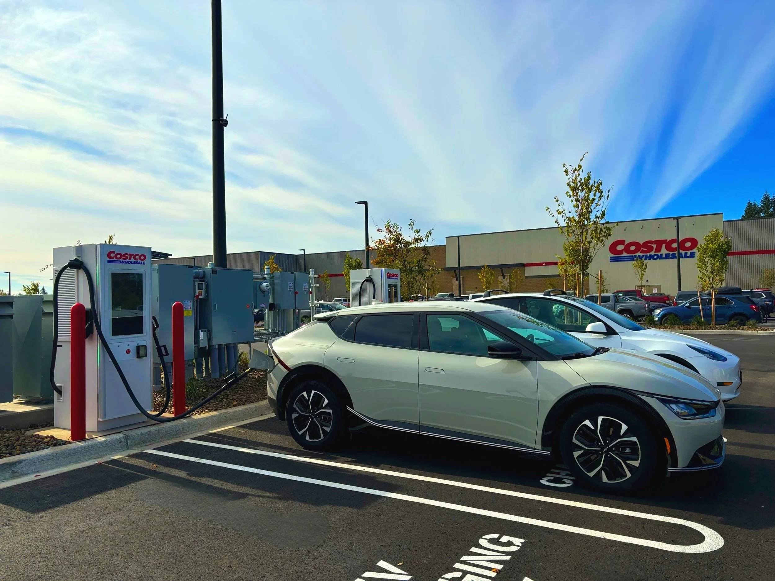 Electric vehicle charging station at Costco parking lot with a white electric car plugged in, and other vehicles in the background.