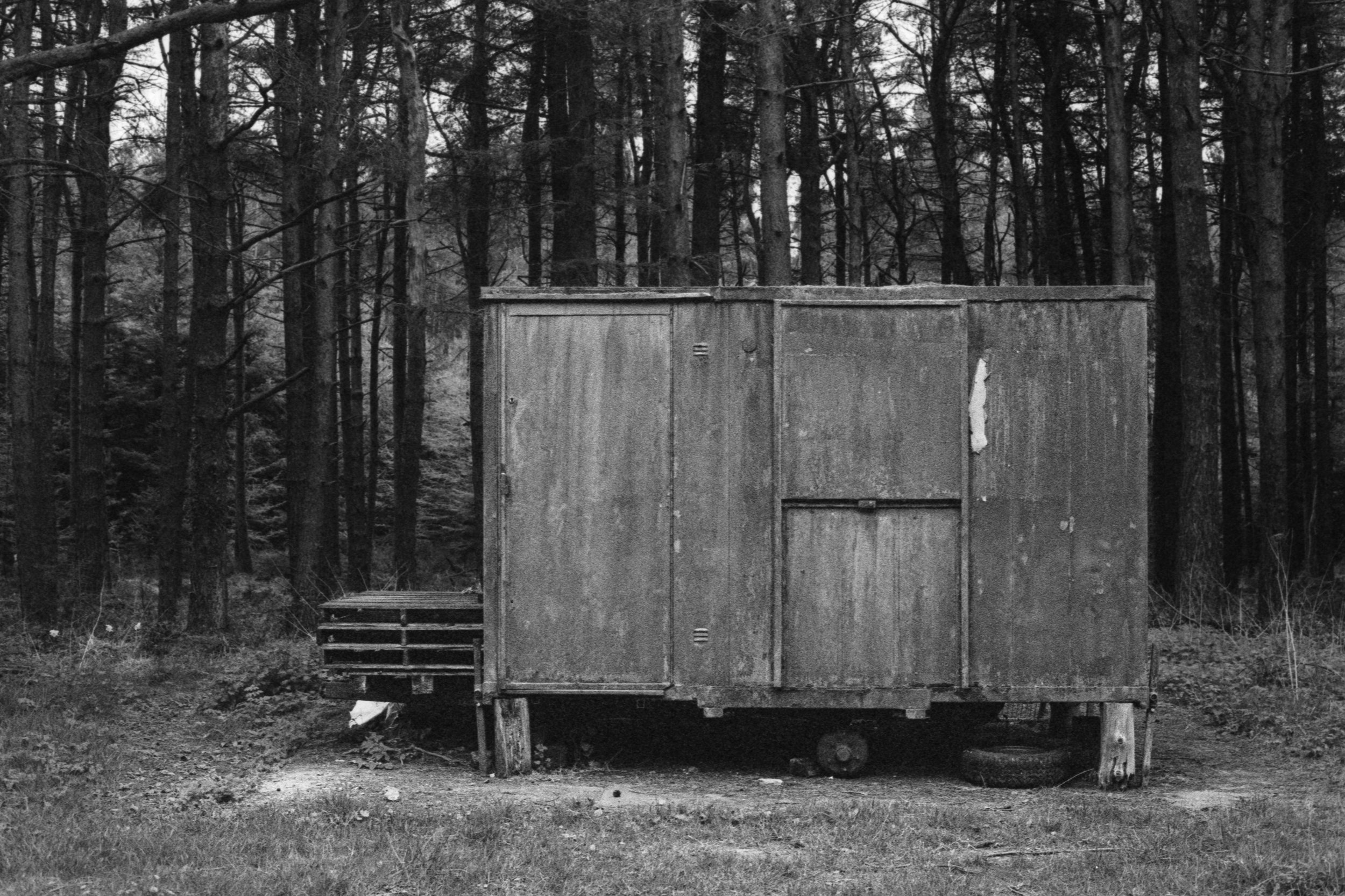 A small, weathered wooden shed in a forest, with a wooden pallet on one side and an upturned tire underneath, in black and white.