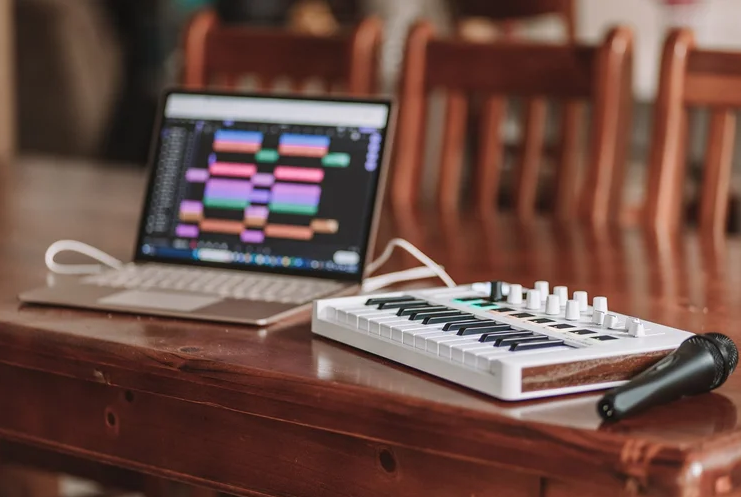 Laptop displaying music production software beside a small MIDI keyboard controller and microphone on a wooden table.