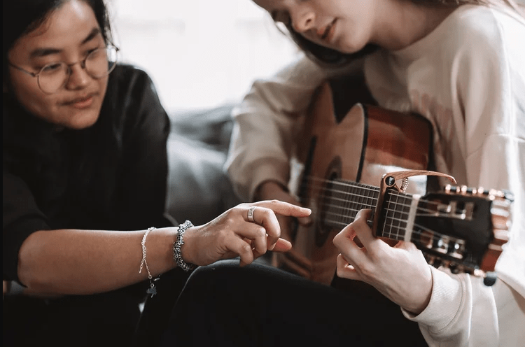 Instructor pointing at a student’s fingers on an acoustic guitar fretboard while guiding them on how to play a chord.