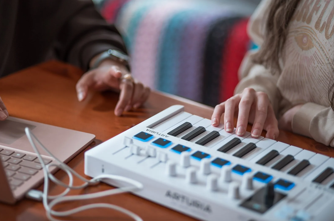 Child playing notes on a small MIDI keyboard controller connected to a laptop while another person gestures nearby.