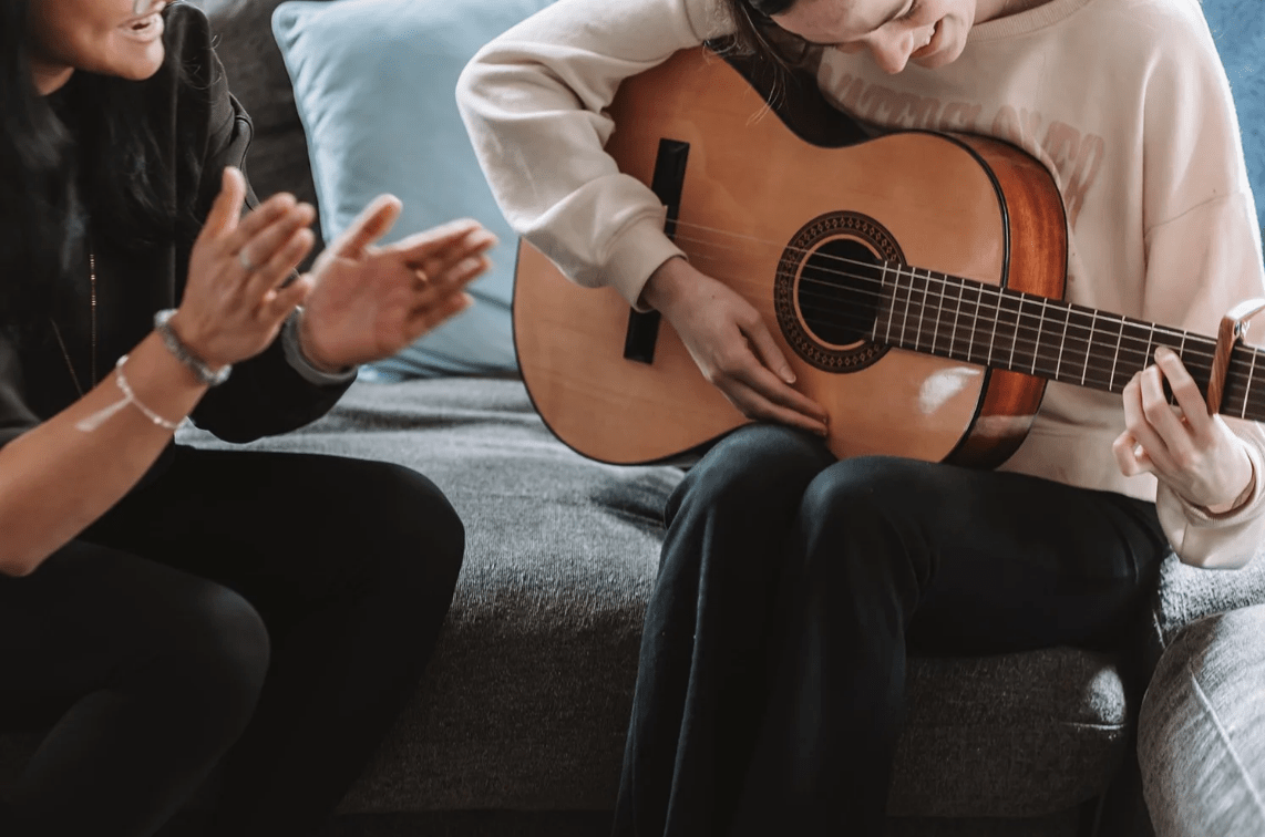 Adult clapping while a child plays an acoustic guitar on a couch during a music lesson.